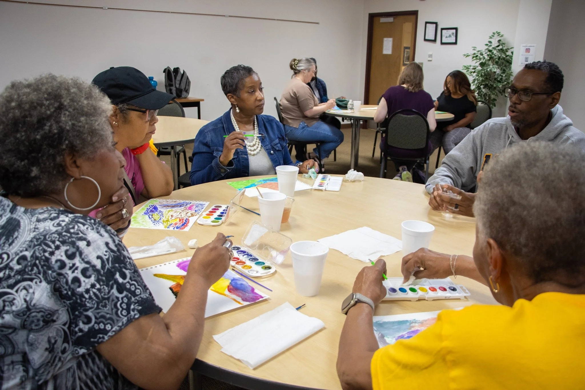 People sitting around a table engaging in a watercolor painting activity, with art supplies including watercolor palettes and paper in a room with neutral-colored walls and framed pictures.