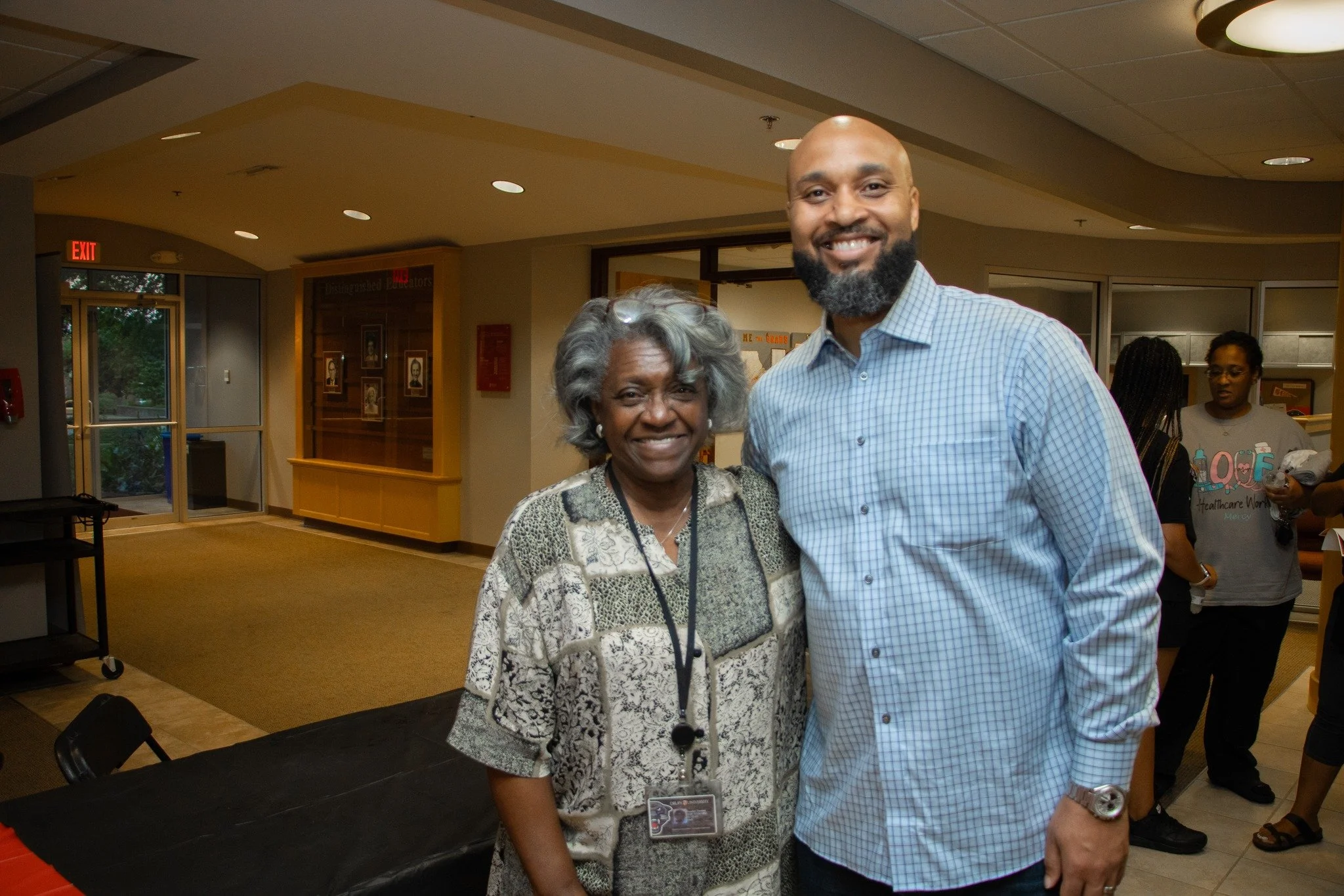 A smiling woman with gray hair and a man with a bald head and beard standing together indoors. The woman wears a patterned blouse and the man is in a checkered shirt, both facing the camera.