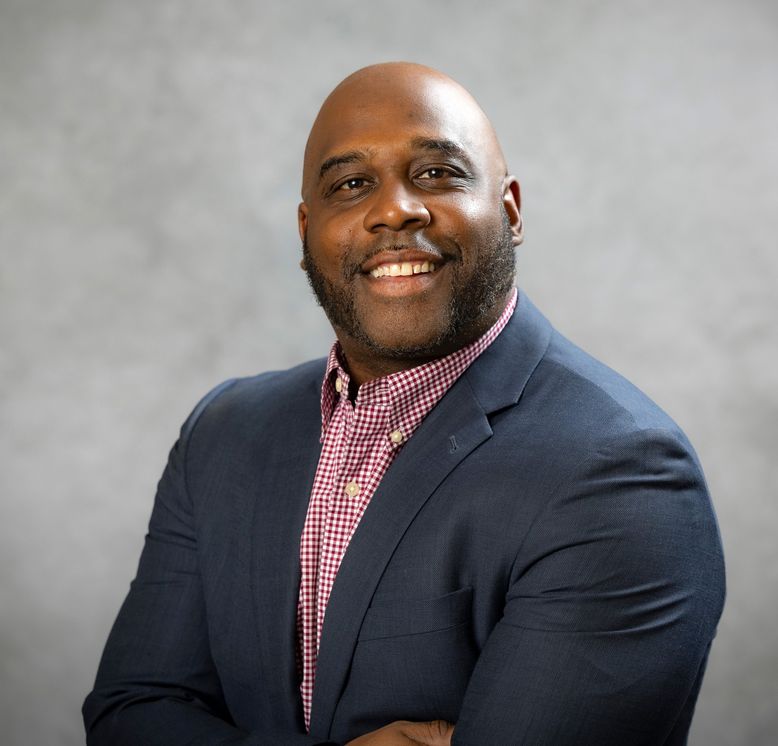 Close-up of a smiling African American man in a navy suit and a red checkered dress shirt, against a gray background.