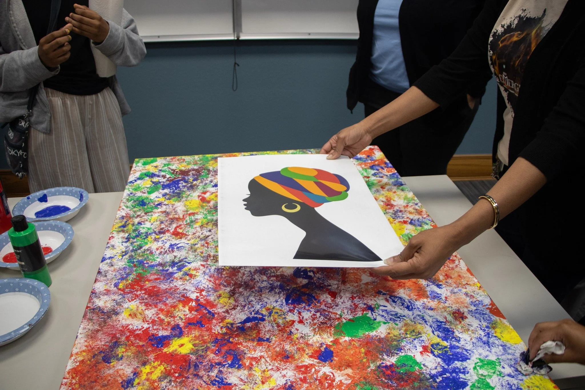 People are holding a printed artwork of a woman's head profile with colorful abstract hair, showing a black silhouette against a splattered multicolored paint background.