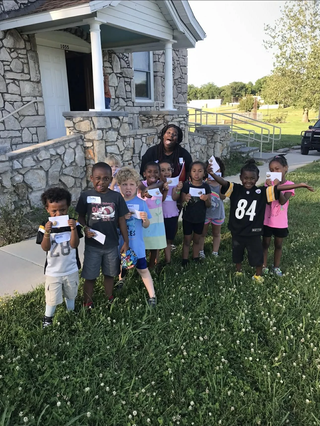 Group of children and a woman standing on a grassy area in front of a stone house, holding papers and smiling.