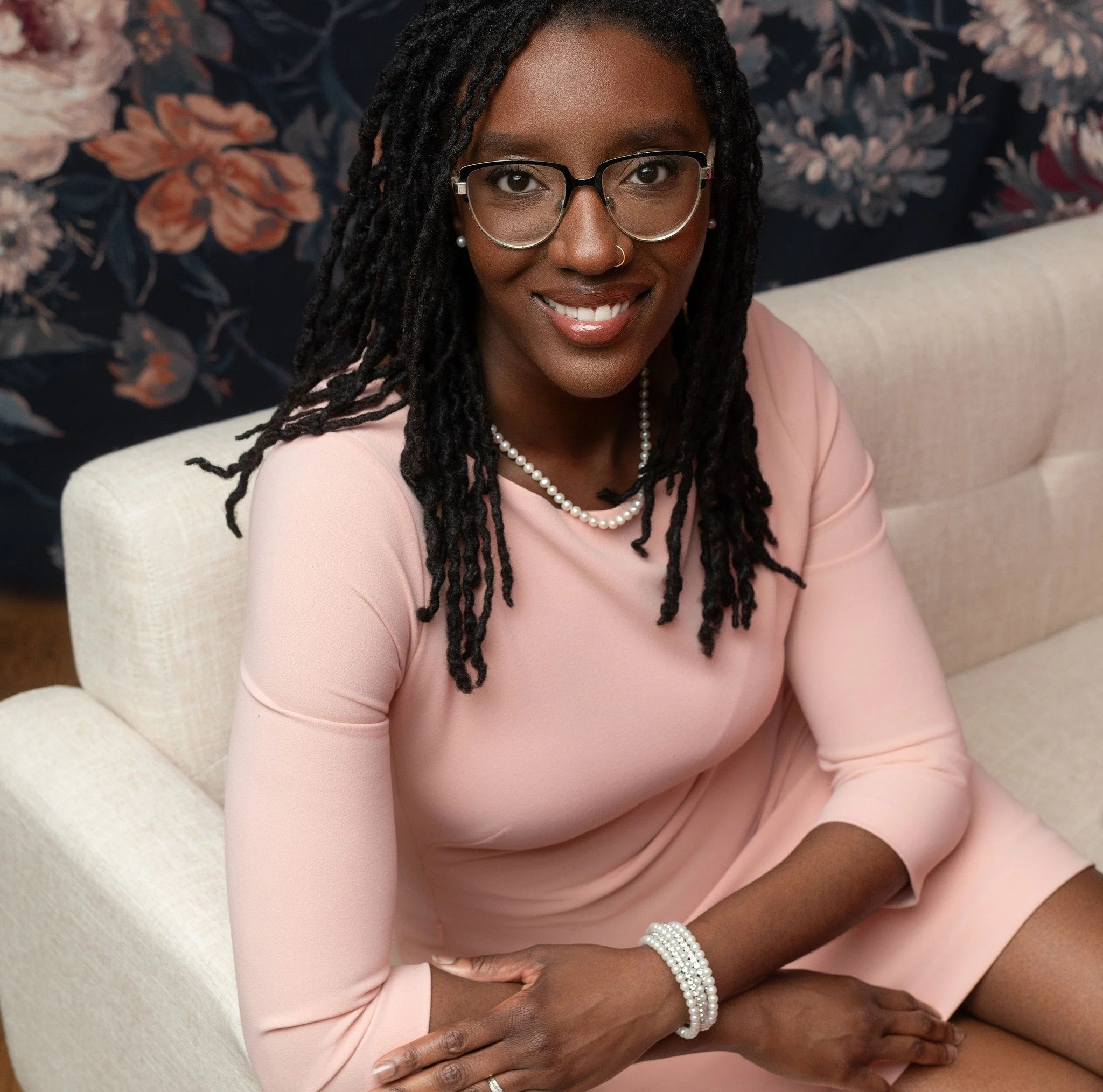 Portrait of a smiling Black woman with glasses and dreadlocks, wearing a pink top and pearl jewelry, sitting on a beige couch with floral wallpaper in the background.