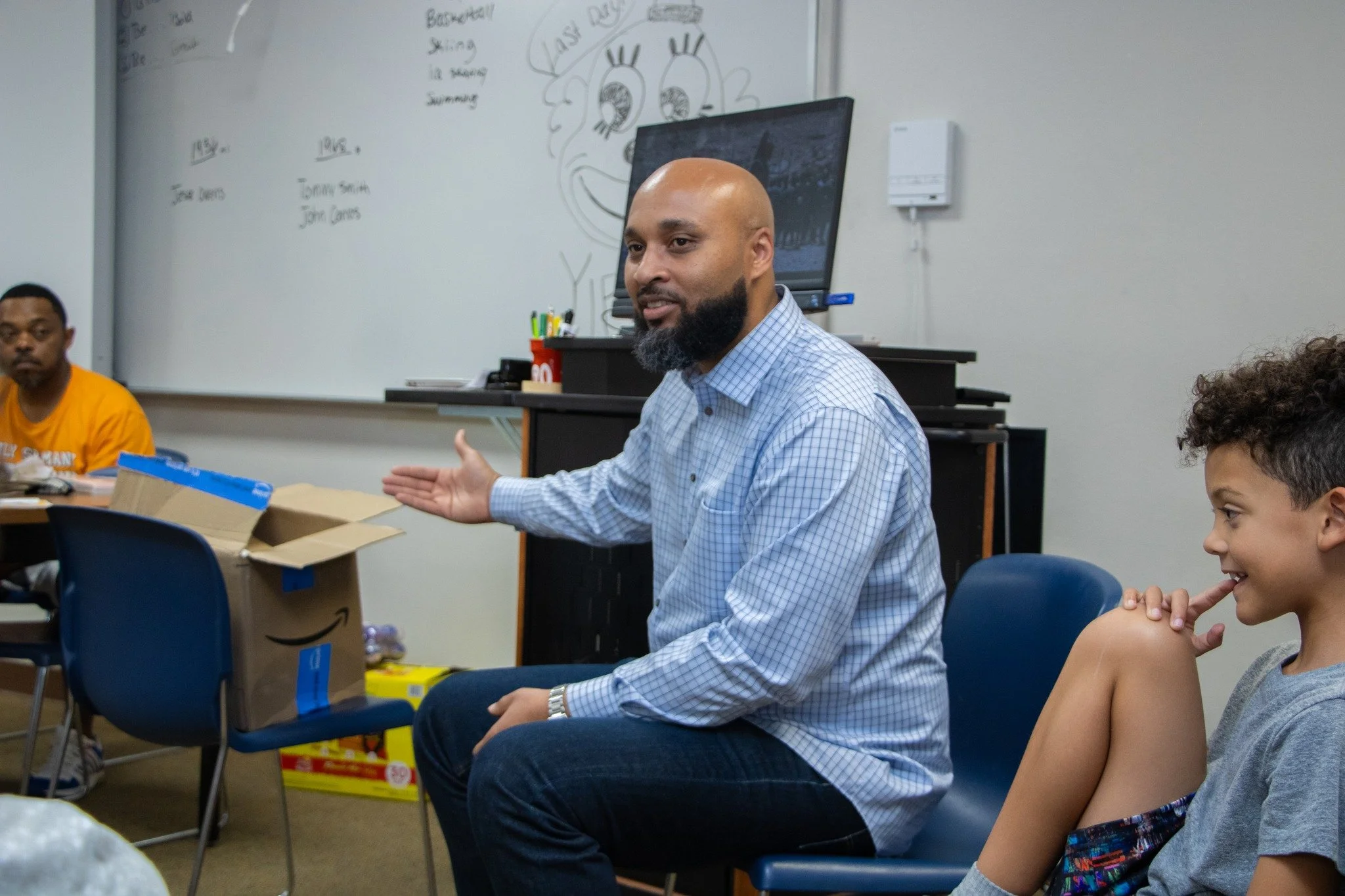 A man with a bald head and beard wearing a blue checkered shirt is speaking, sitting at a table with children and other people in a classroom setting. There is a whiteboard with writing and drawings, including a large cartoonish face with exaggerated