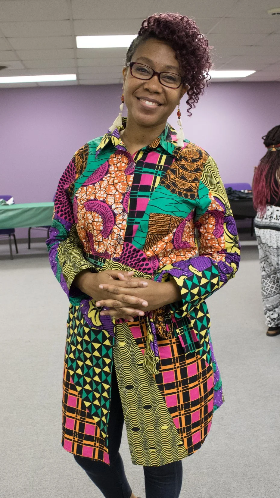 A woman with glasses and purple curly hair, smiling, wearing a colorful patchwork dress with African prints, standing indoors with a purple wall in the background.