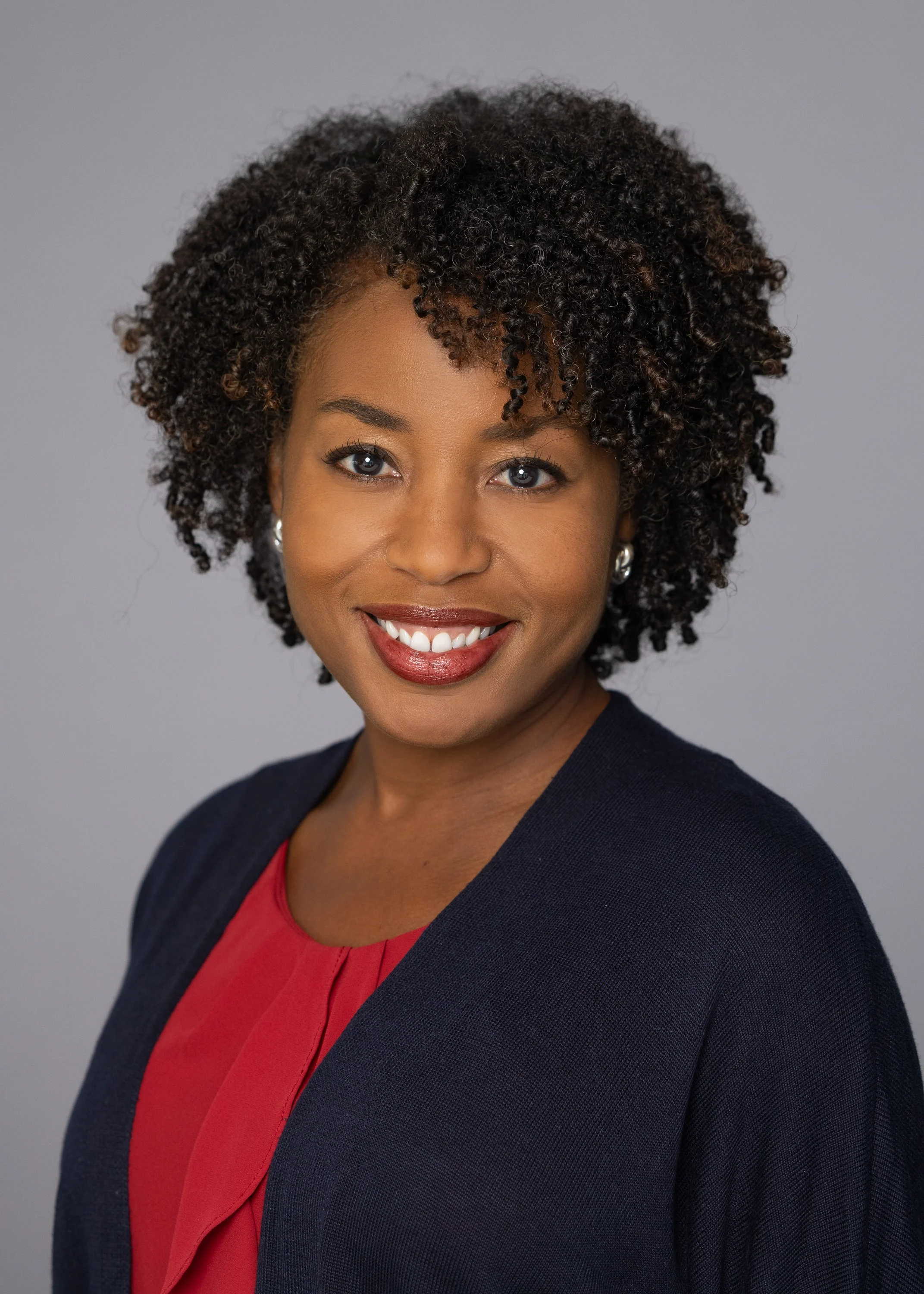 Close-up of a woman with dark curly hair and blue eyes smiling, wearing a red top and a dark blazer, against a plain gray background.