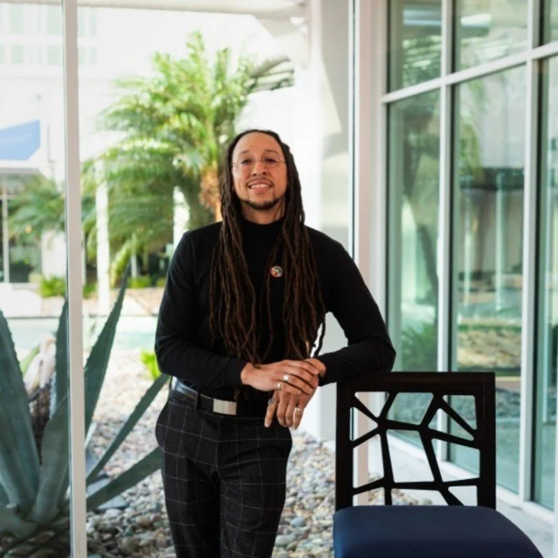 A smiling person with long dreadlocks and glasses standing indoors in front of large windows with plants outside.