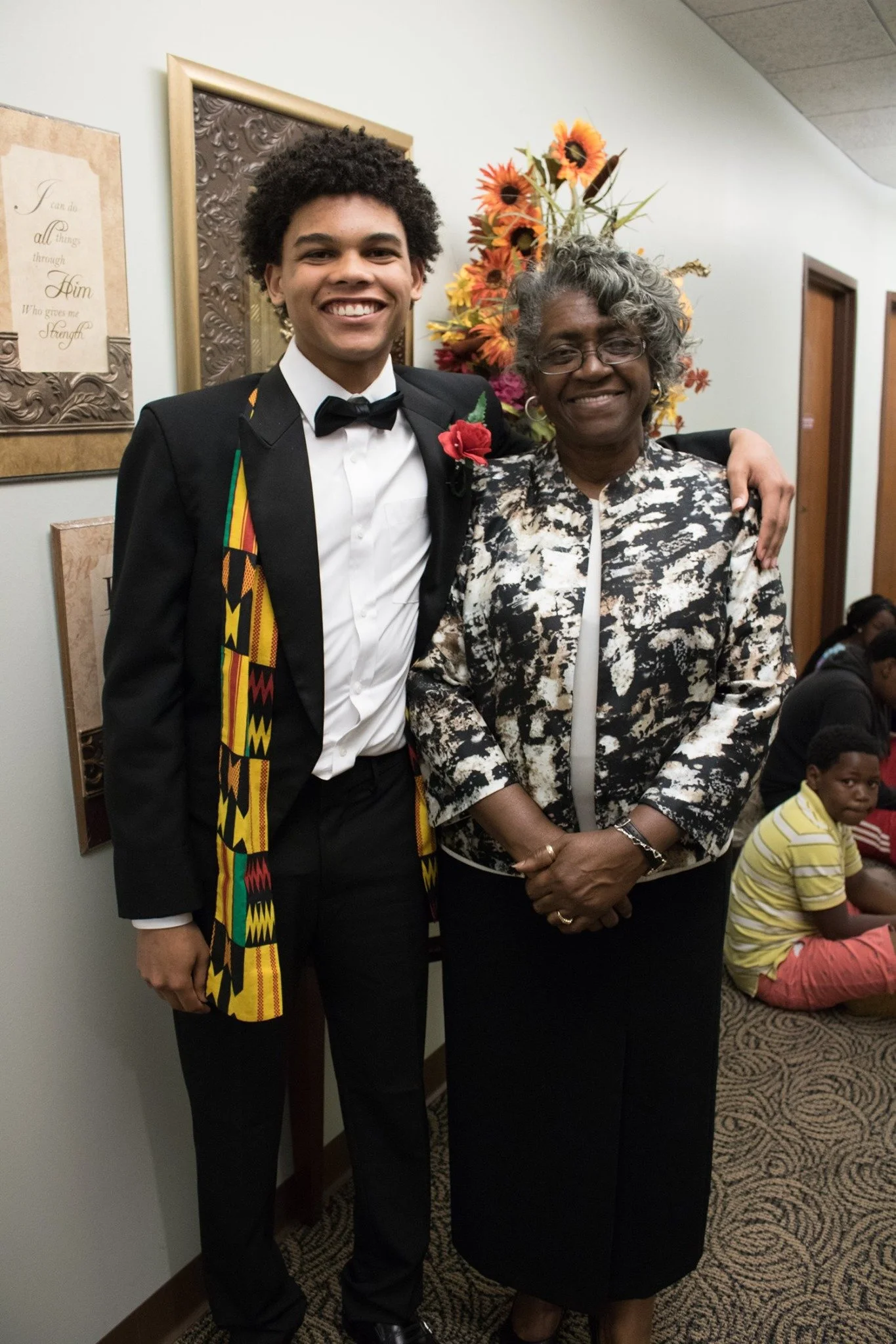 A young man in a tuxedo with a colorful kente stole, standing next to an older woman in a patterned black and white blazer, both smiling, with flowers in the background, in an indoor setting.