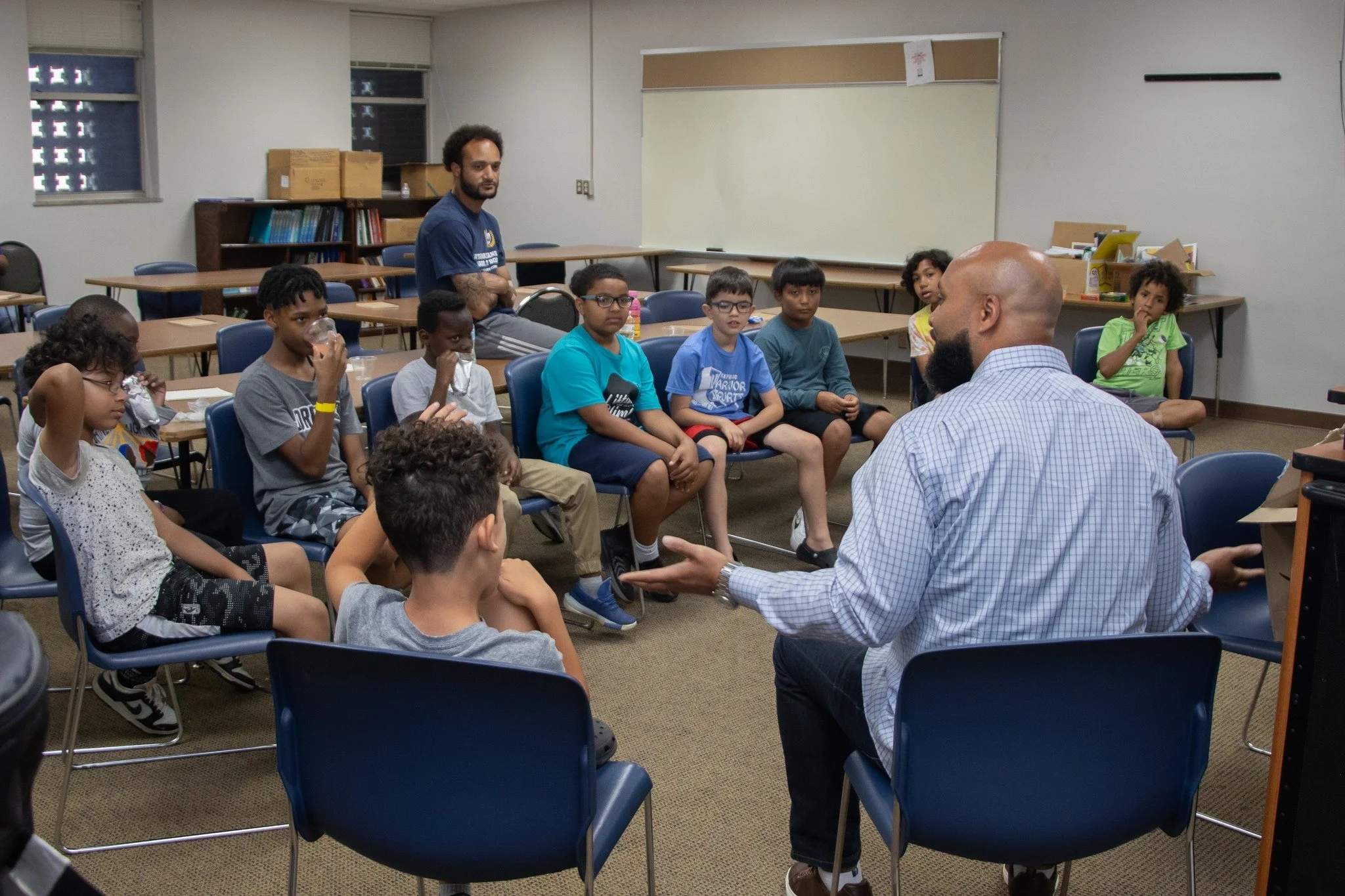 A group of children sitting around a table in a classroom, attentively listening to a man with a beard who is speaking to them, while another man with a beard and glasses stands nearby. The classroom has shelves with books and boxes, whiteboards, and