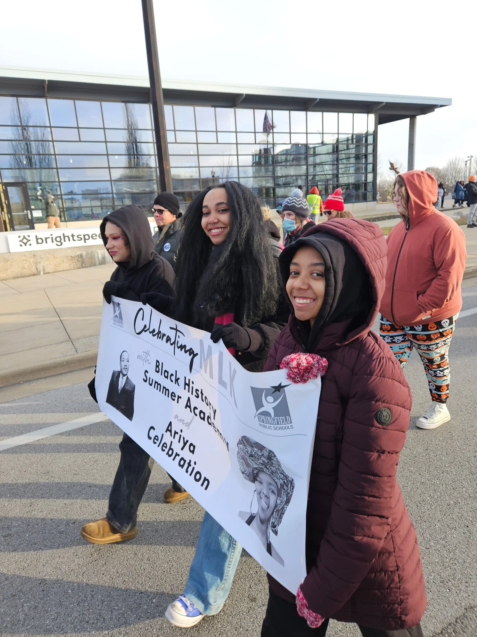 Young people participating in a Black History Month parade outside a modern building, carrying a banner that references a summer academic program and celebration.