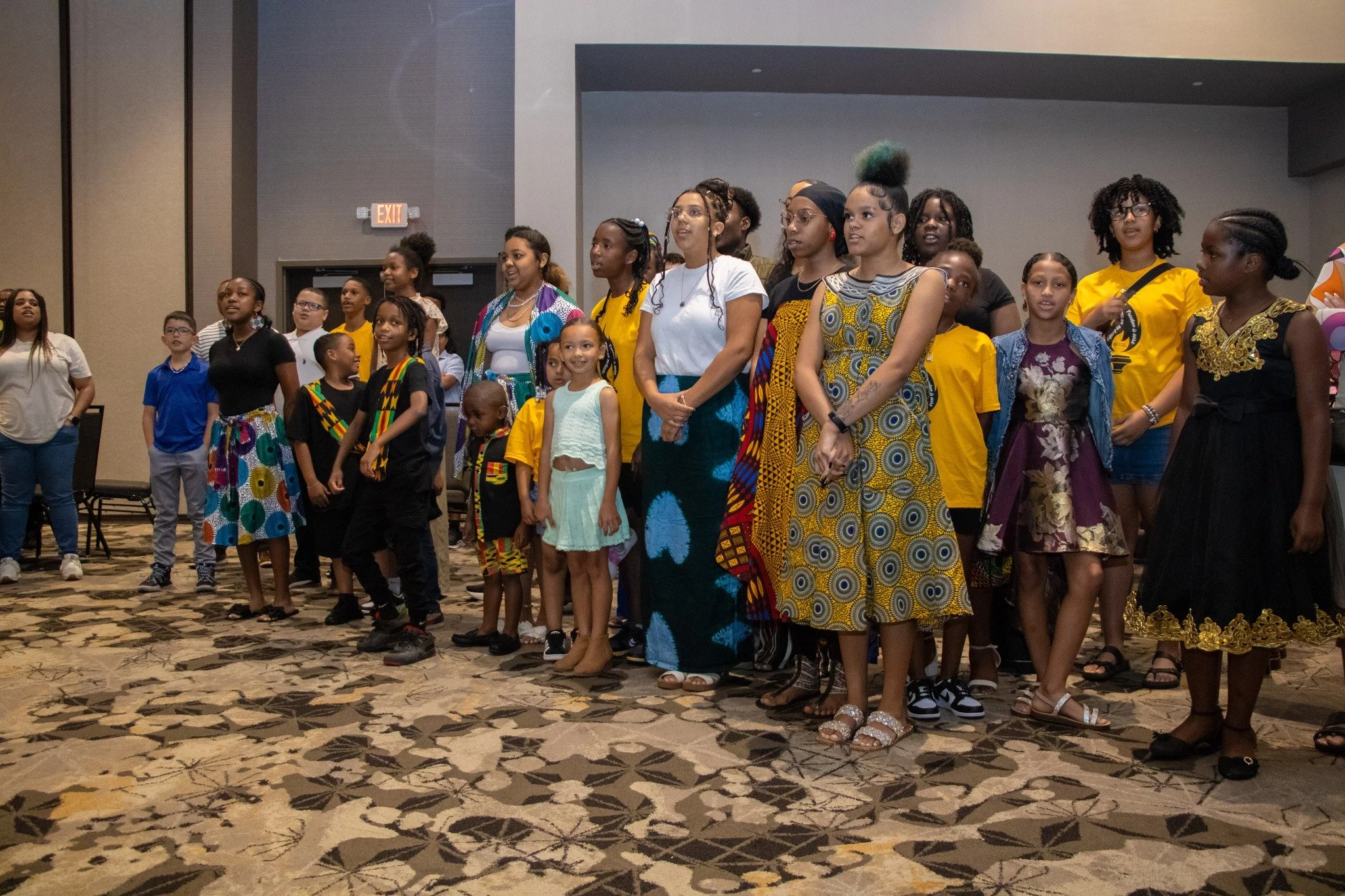 A group of diverse women and girls standing together in a conference room, some wearing colorful traditional African dresses, others in casual clothing, facing forward and listening attentively.