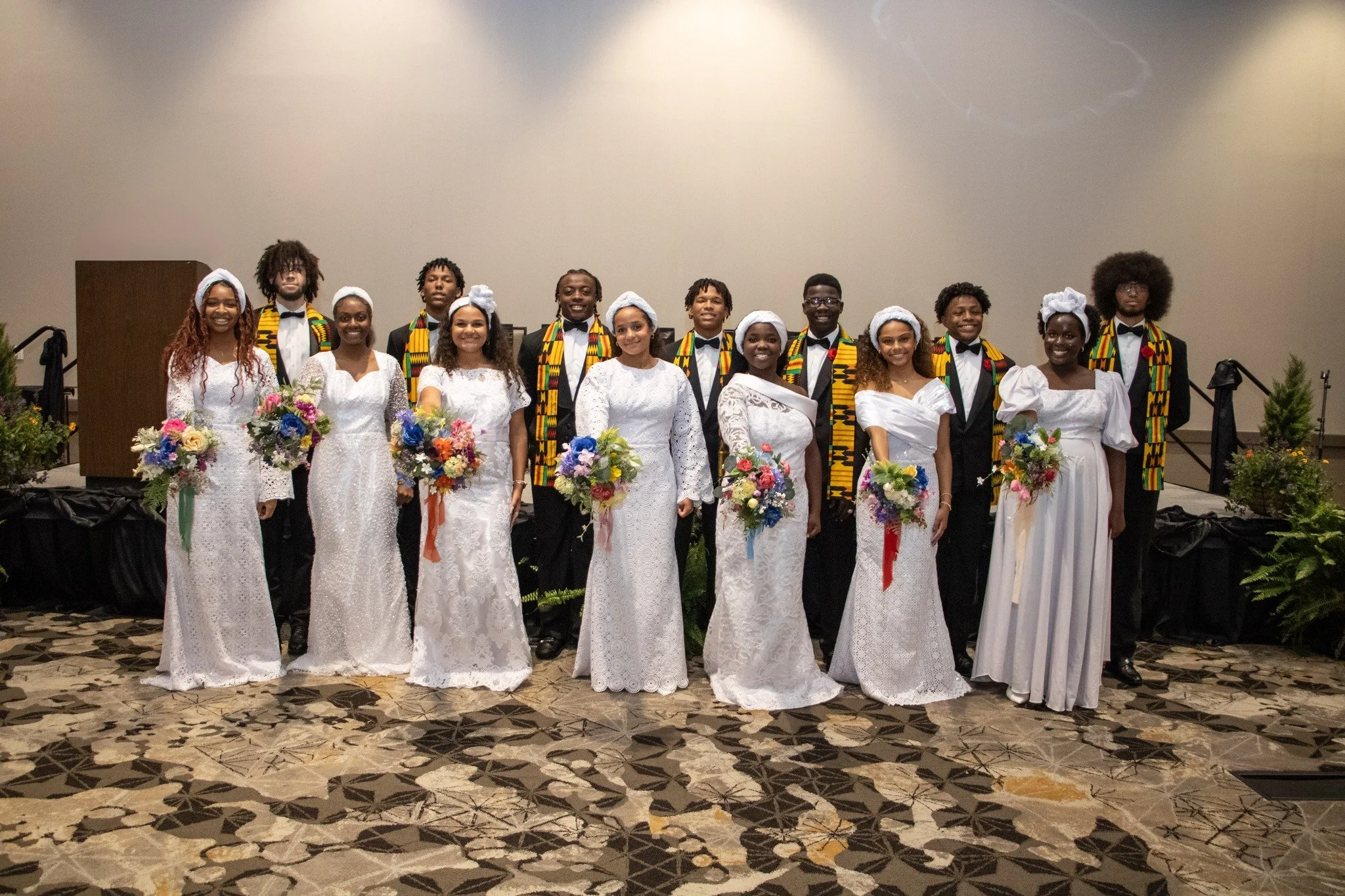 Group of young people at a formal event, dressed in white dresses and black suits with colorful kente cloth scarves, holding bouquets of flowers, standing together on a patterned carpeted floor against a plain background.