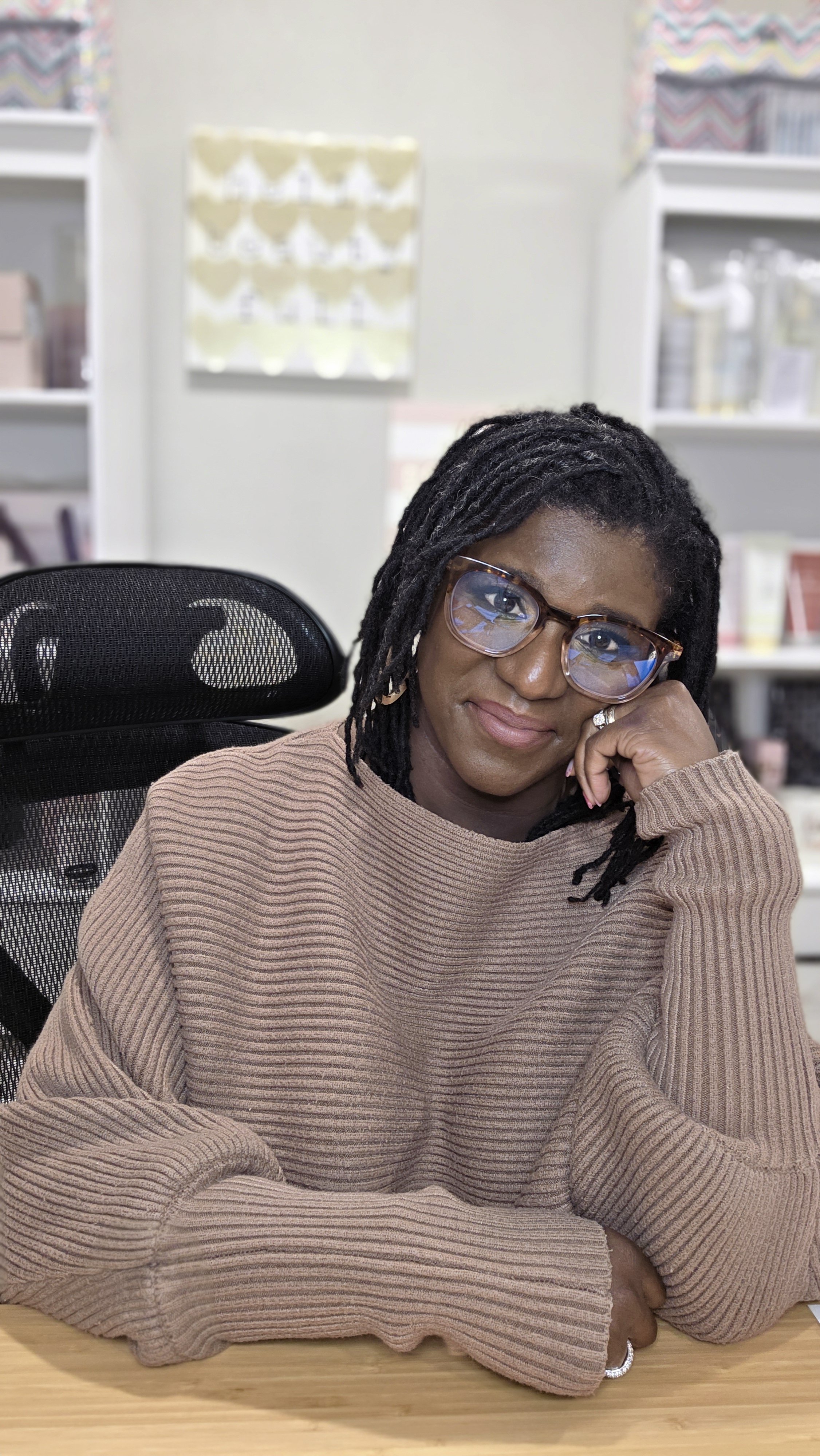A woman with glasses and dreadlocks sitting at a desk, smiling and resting her head on her hand.