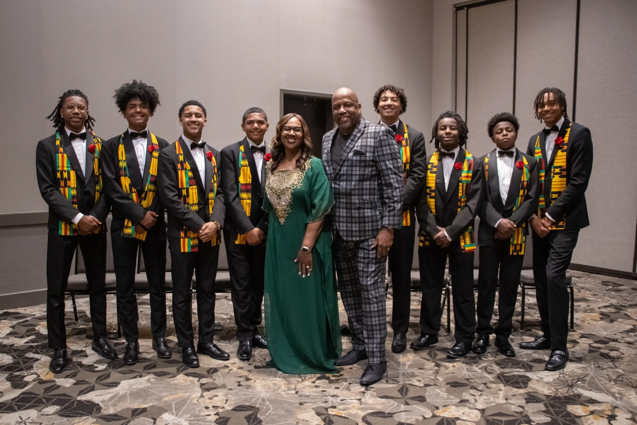 Group of nine young men in tuxedos with kente cloth stoles, two adults in the center, one woman in a green dress, in an indoor setting.