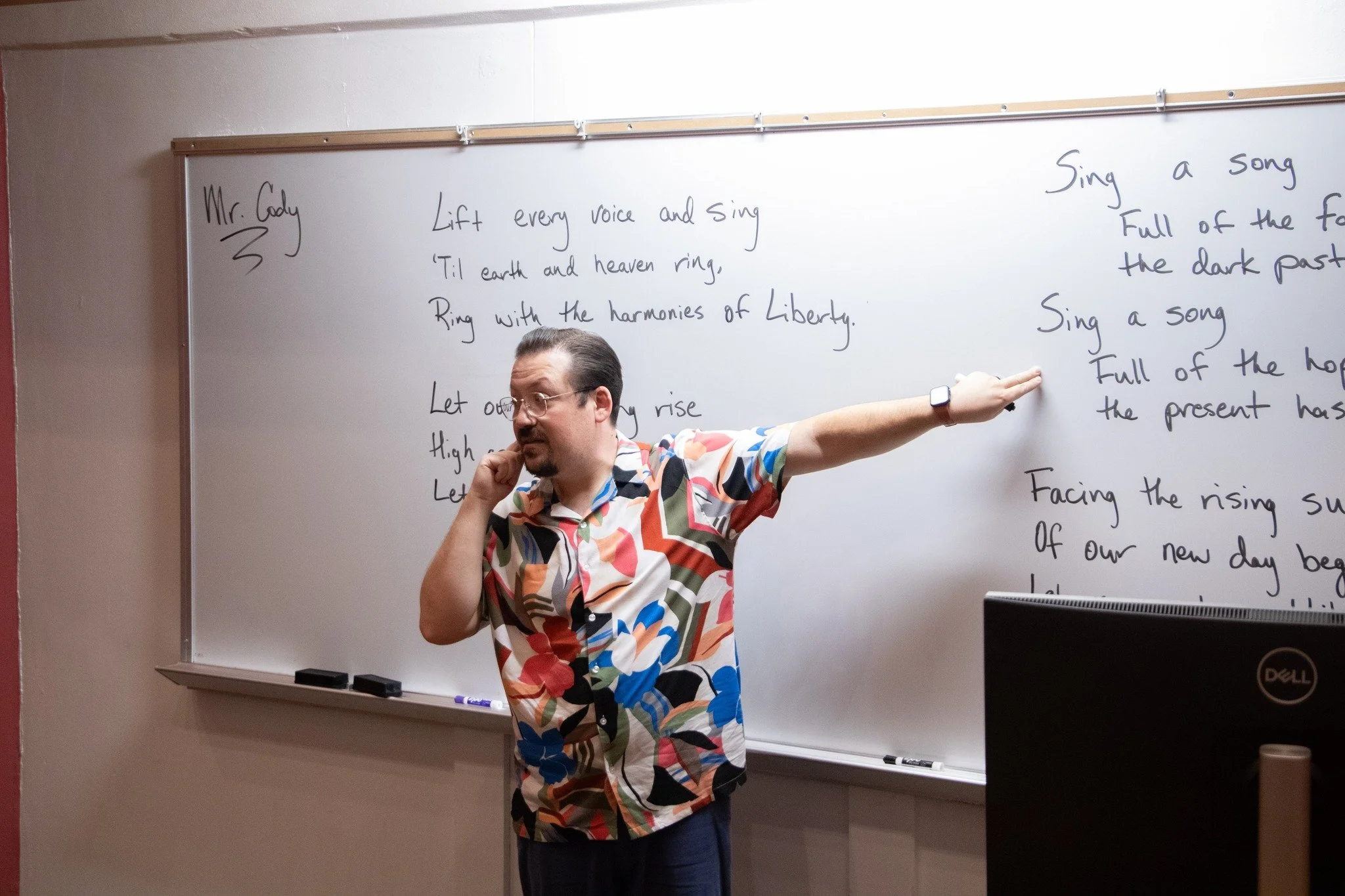 A man wearing a multicolored, patterned shirt standing in front of a whiteboard with handwritten song lyrics, pointing at the whiteboard with his right hand and resting his left hand on his cheek.