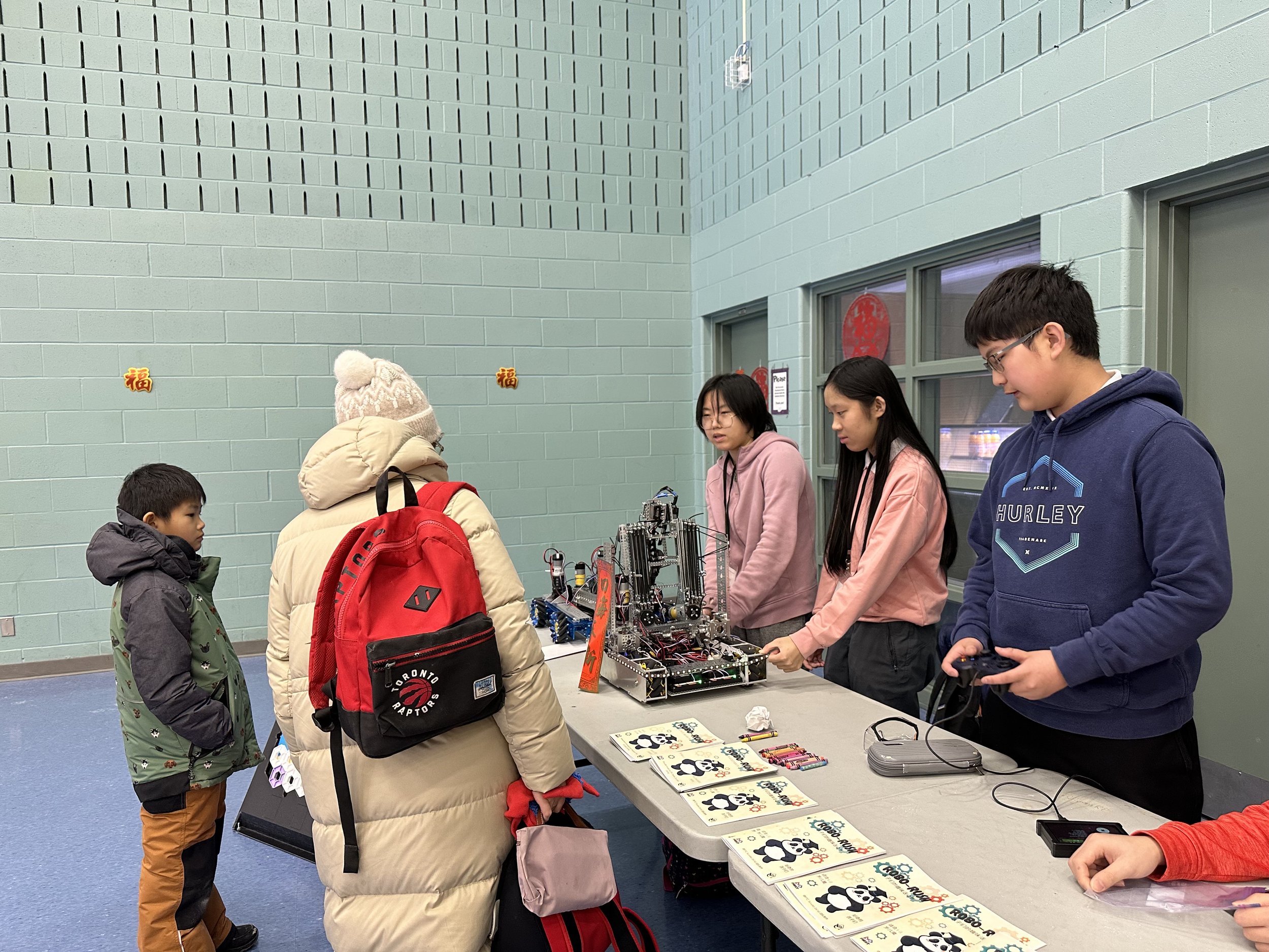 People at a robotics demonstration table with pamphlets, robots, and gaming controllers in a community center.