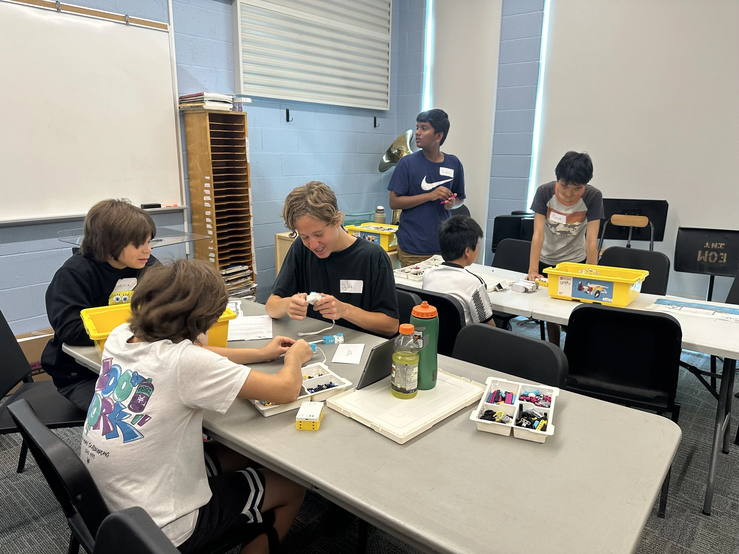 A group of children and teenagers working on building and programming Lego robotics in a classroom setting, with tables, bins of Lego pieces, and a whiteboard.