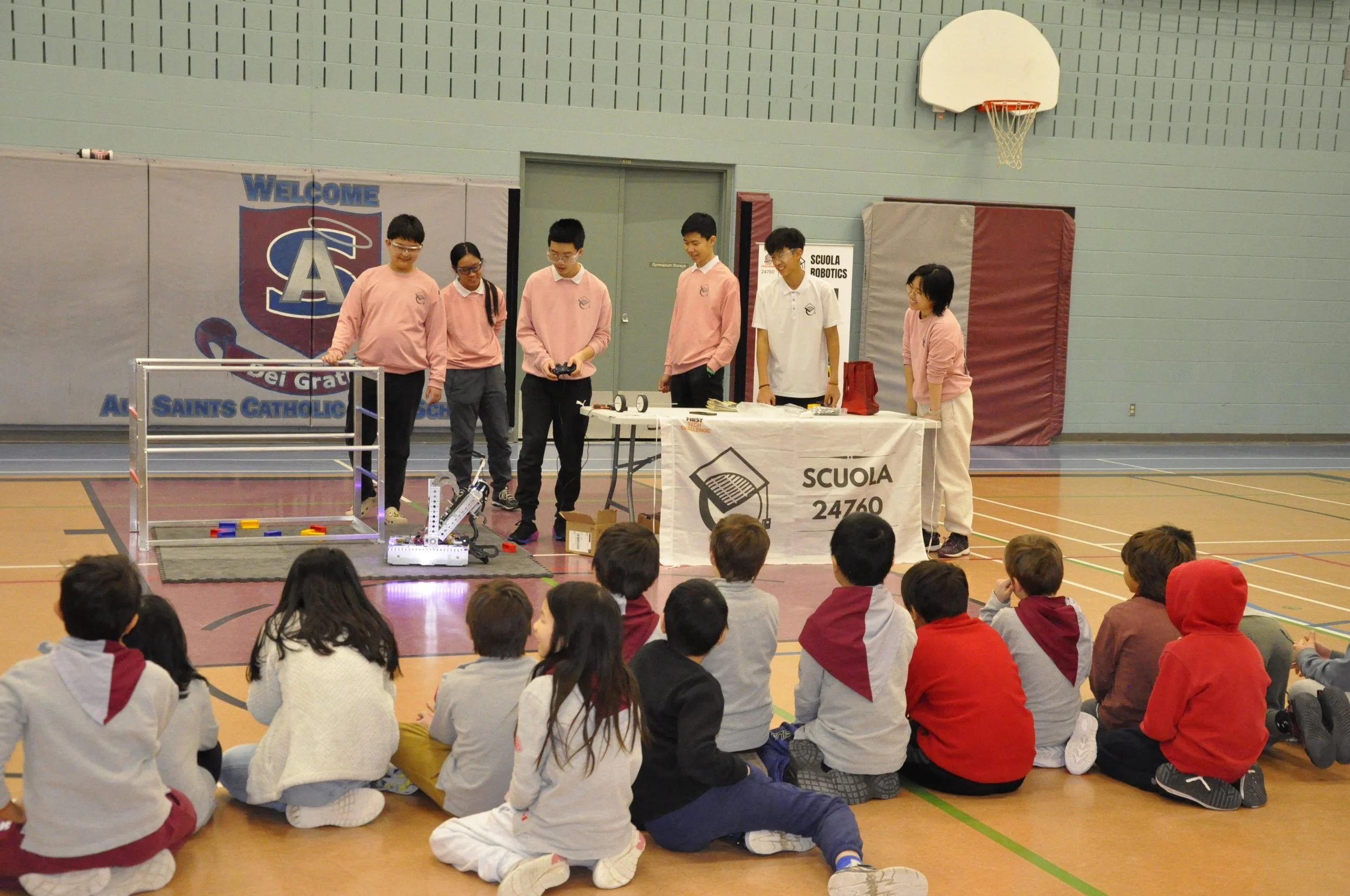 Students demonstrating robotics projects to younger students in a gymnasium, with a banner reading 'SAINTS CATHOLIC' in the background and a table with various robotic equipment.