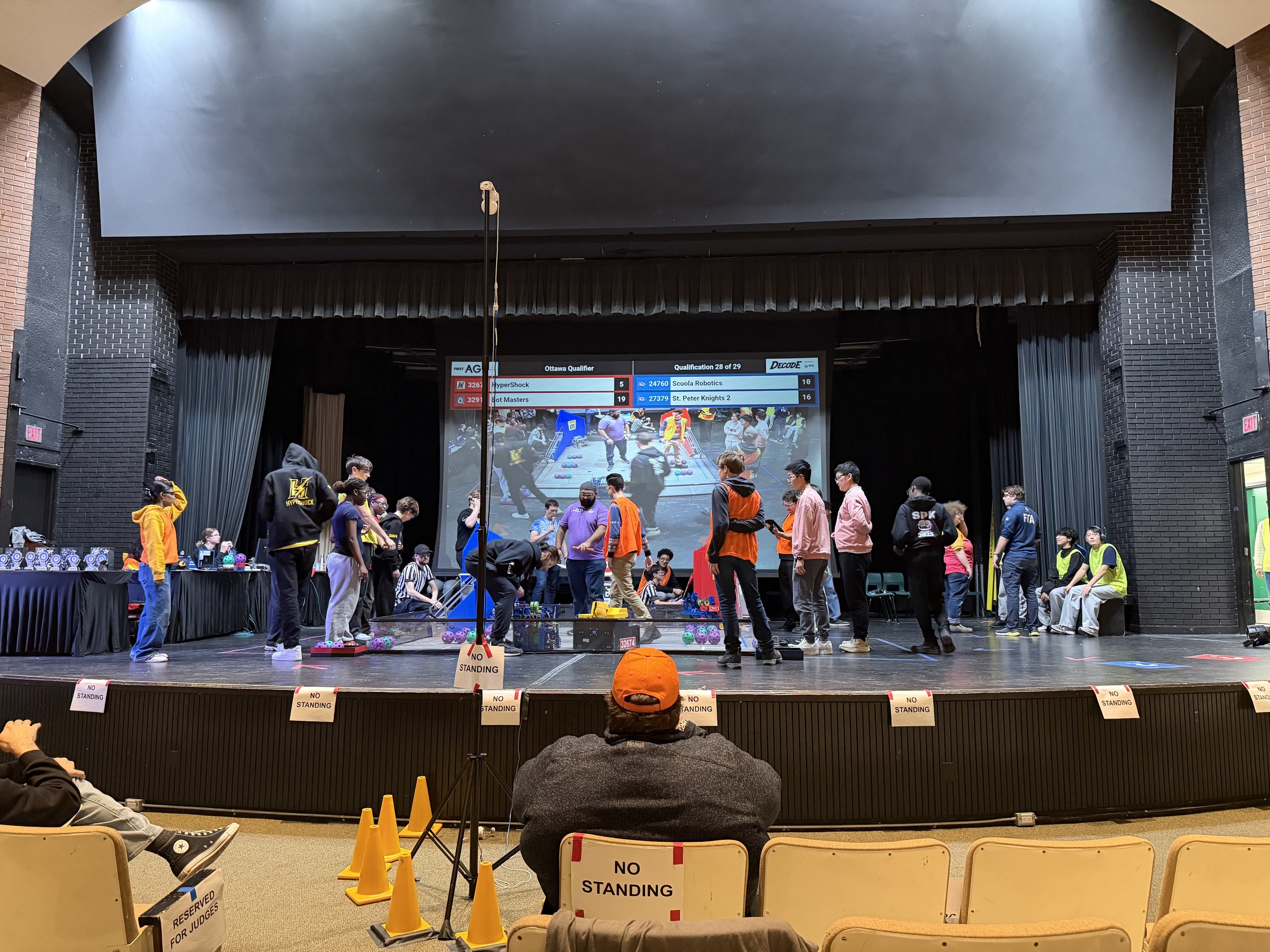 A robotics competition taking place in an auditorium with a large digital scoreboard showing team names and scores. Several people are on stage, some wearing yellow, orange, or black hoodies, indicating team members or organizers. There are signs on the barricades at the front reading 'NO STANDING' and 'RESERVED FOR JUDGES.' The audience is seated, watching the event, with some individuals taking photos or videos.