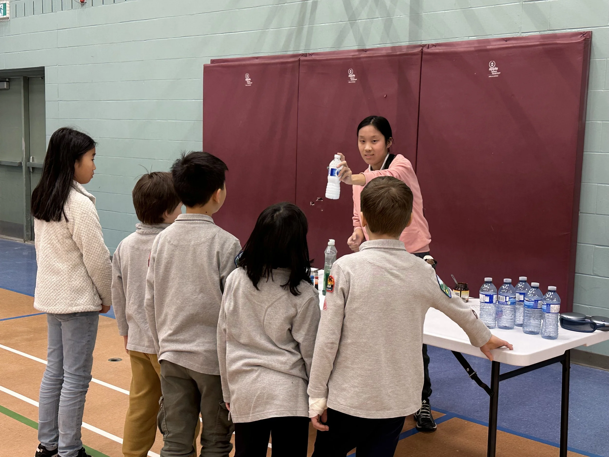 A group of children gathered around a table with water bottles, listening to a girl holding a water bottle in a gymnasium.