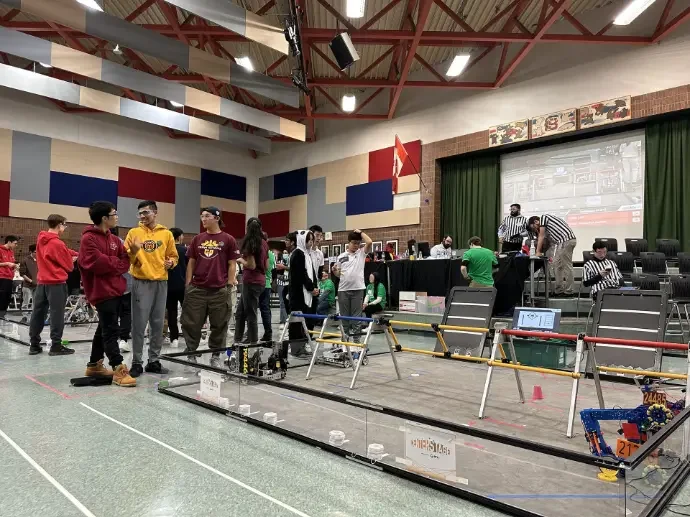 Students and staff in a gymnasium preparing for a robotics competition, with robots and equipment on the floor and a large screen showing projections.