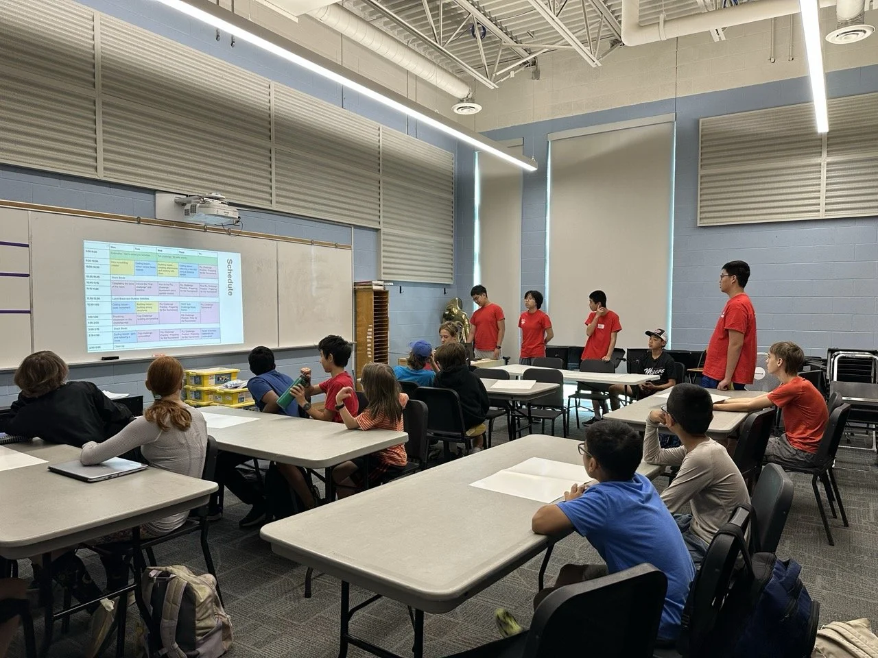 Students in a classroom watching a presentation at the whiteboard, with some students sitting at tables and others standing near the whiteboard.