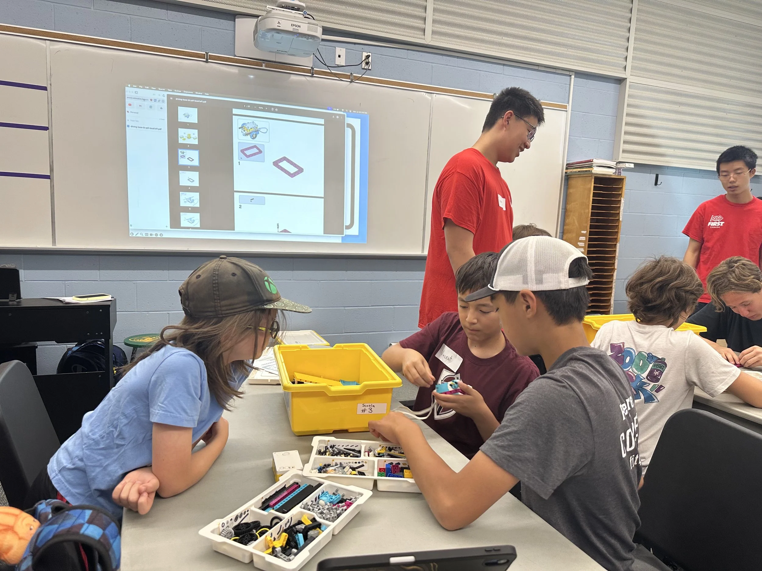 Group of children working on a robotics project in a classroom with teachers supervising. The children are assembling parts from yellow bins, with a whiteboard and projected screen in the background showing instructions.