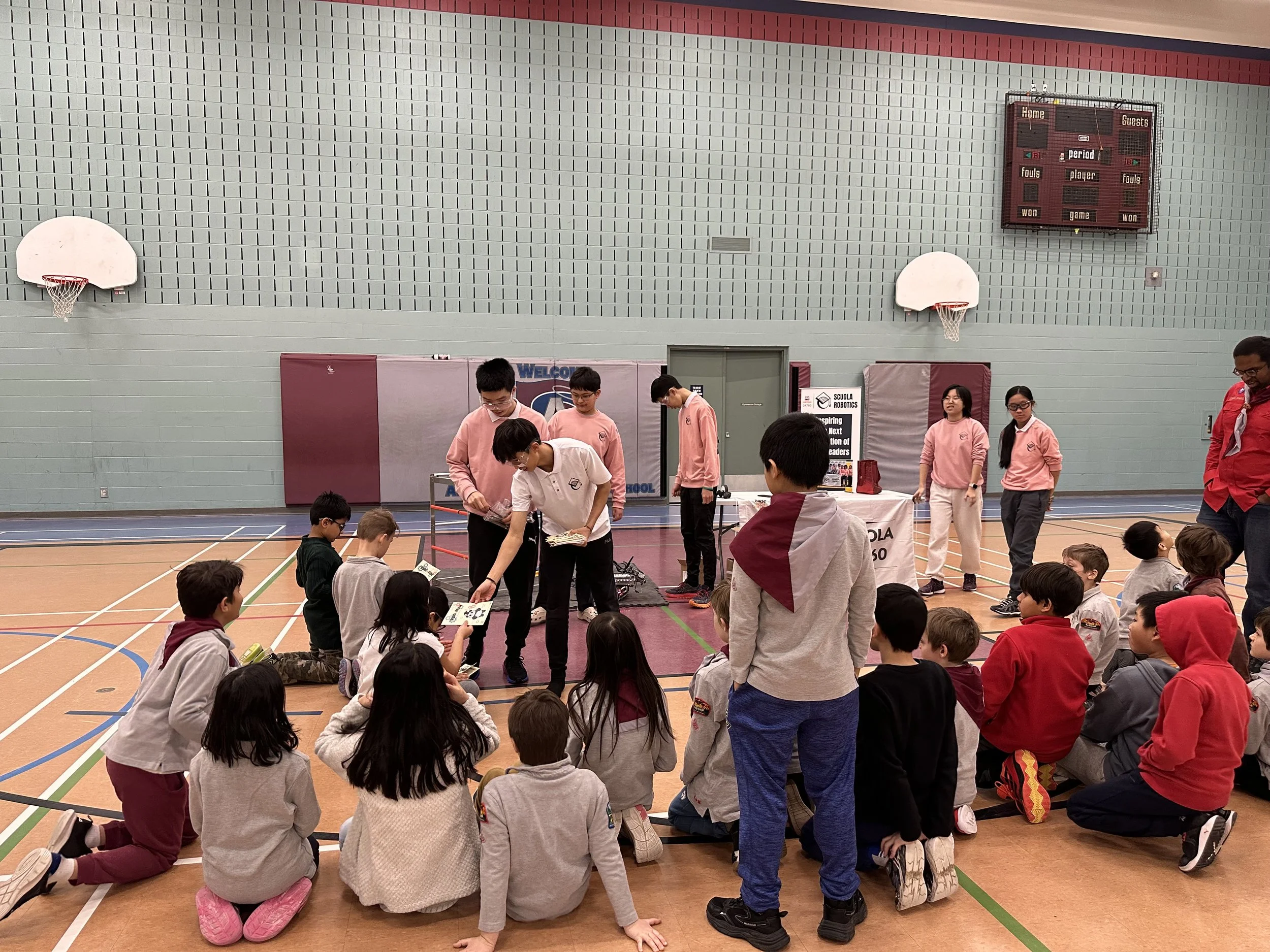 Students participating in a robotics event inside a gymnasium, with some seated on the floor and others standing near tables.