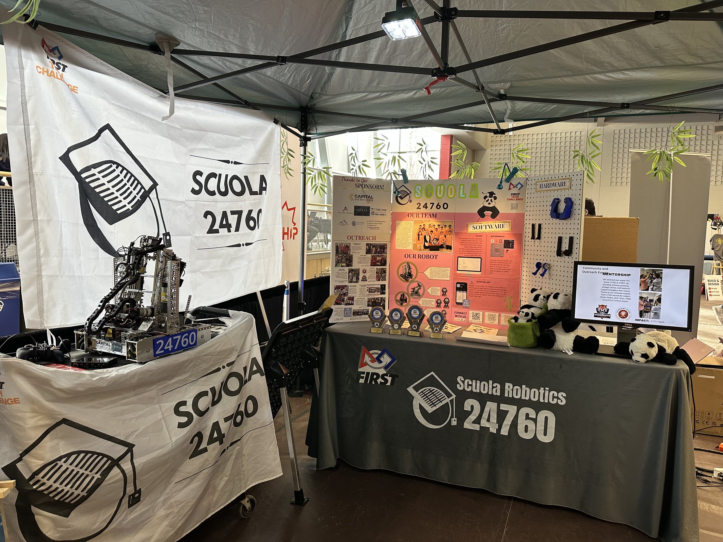 An indoor display booth for Scuola Robotics, featuring banners with the team number 24760, robot models, trophies, & informational posters, under a green canopy with hanging decorative bamboo leaves.