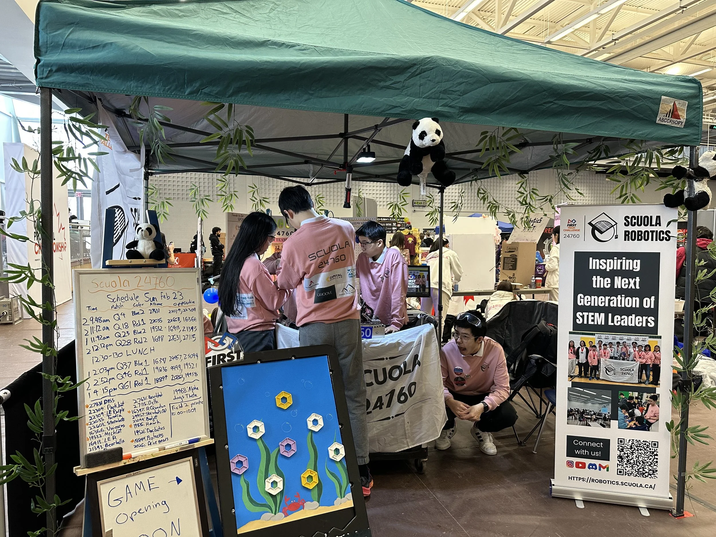 Students and staff at a booth for Scuola Robotics at a STEM event, with signs, panda plush toys, and electronic equipment on display inside a large indoor space.