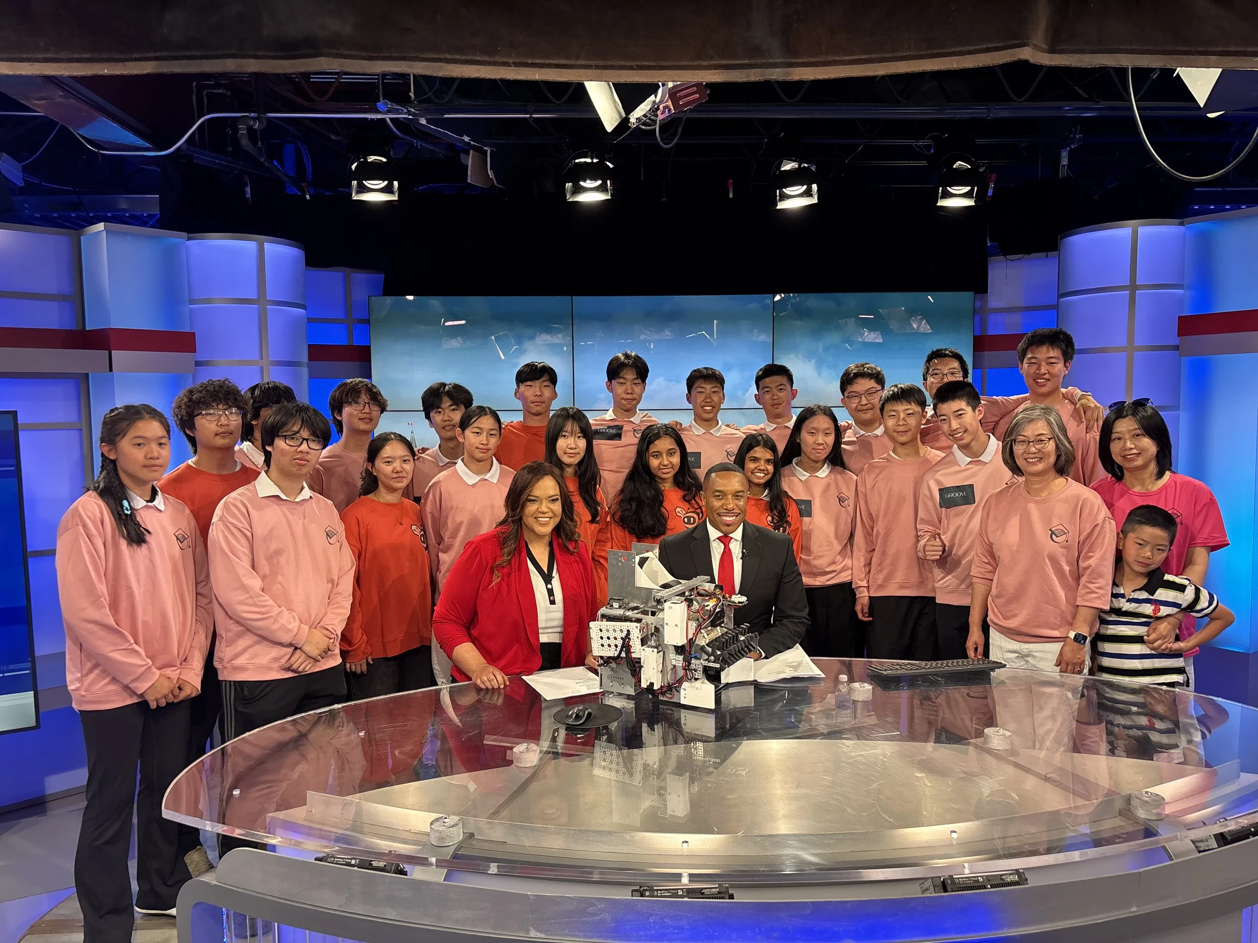 Group of young students and adults in a television studio, posing for a photo behind a glass table with robotics equipment.