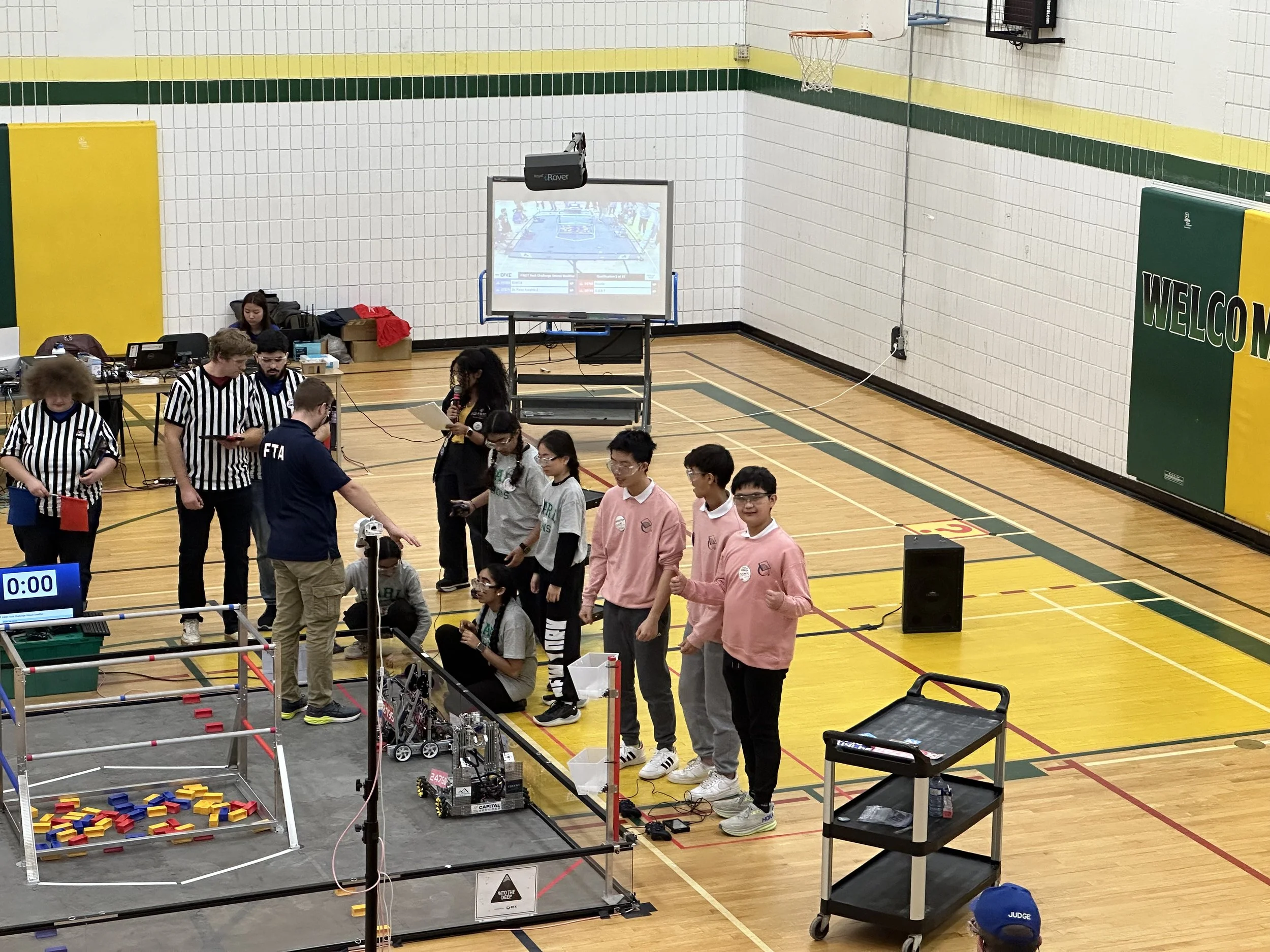 Students and judges participating in a robotics competition inside a gymnasium, with a robot on the floor and a large screen displaying game information.