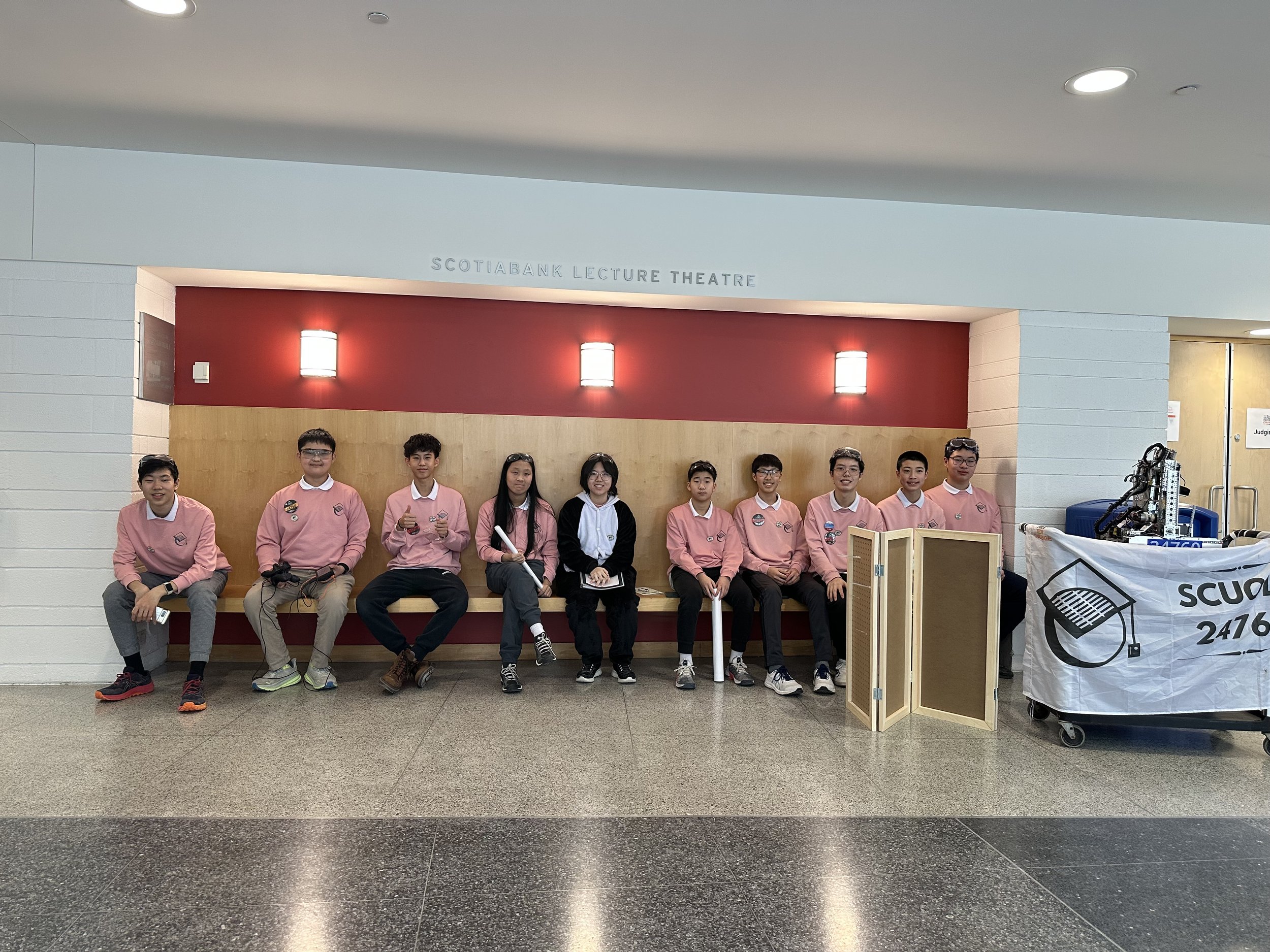 Group of students sitting on a wooden bench in front of red and beige wall at Scotiabank Lecture Theatre, some wearing pink uniforms, some holding papers, next to a robot with a cart, in an indoor setting.