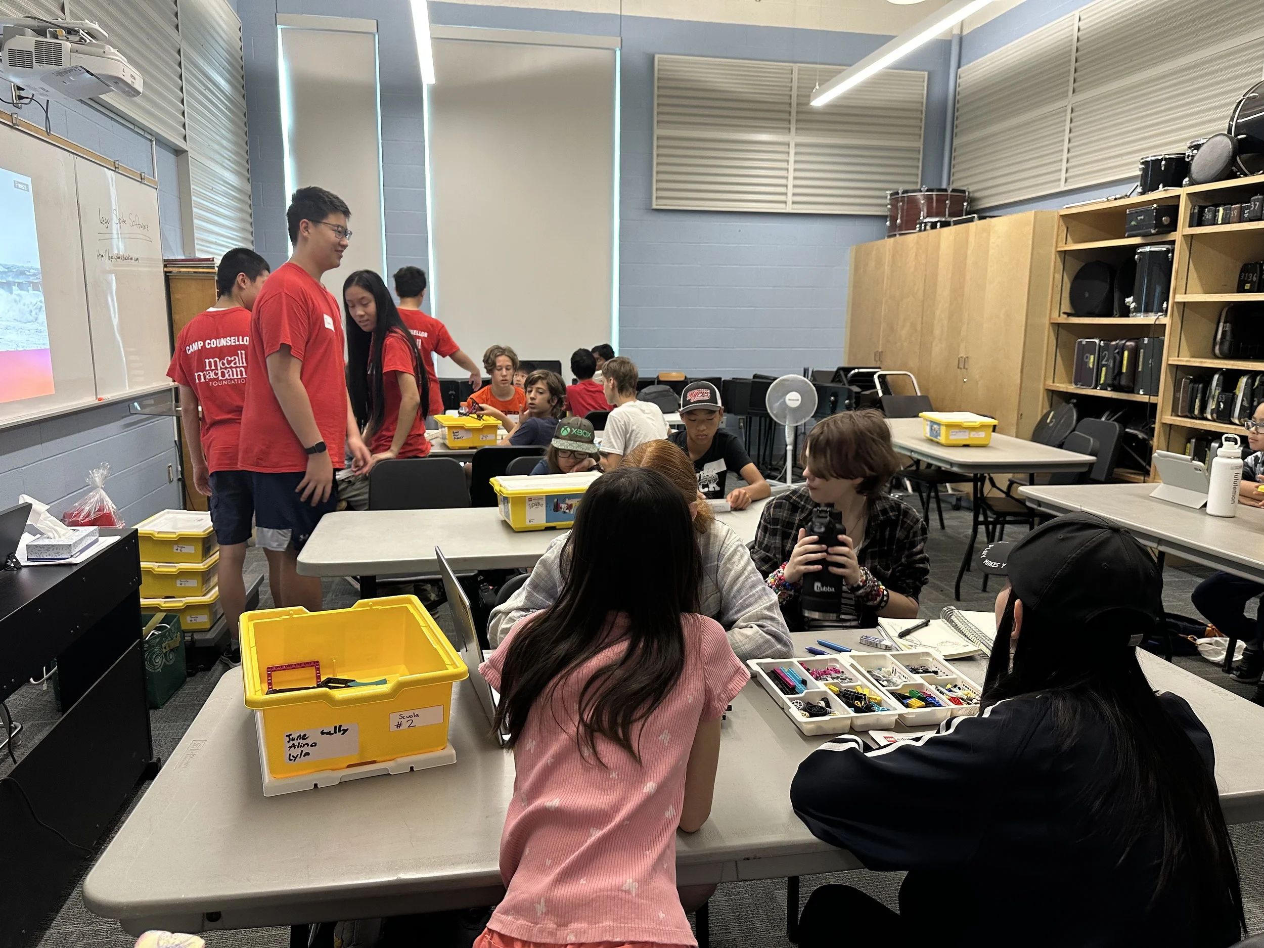 Classroom filled with students engaging in a hands-on activity with various supplies and LEGO pieces, some students are standing near the whiteboard, while others are seated at tables working with LEGO brick kits.