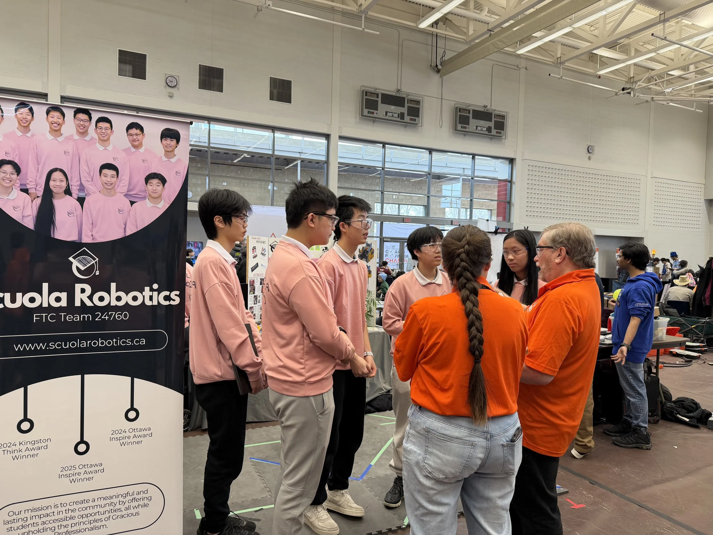 A group of students in pink sweatshirts standing in a row at an indoor robotics event, engaging in conversation with an adult in orange. Behind them, there are display boards and a large banner for a robotics team.
