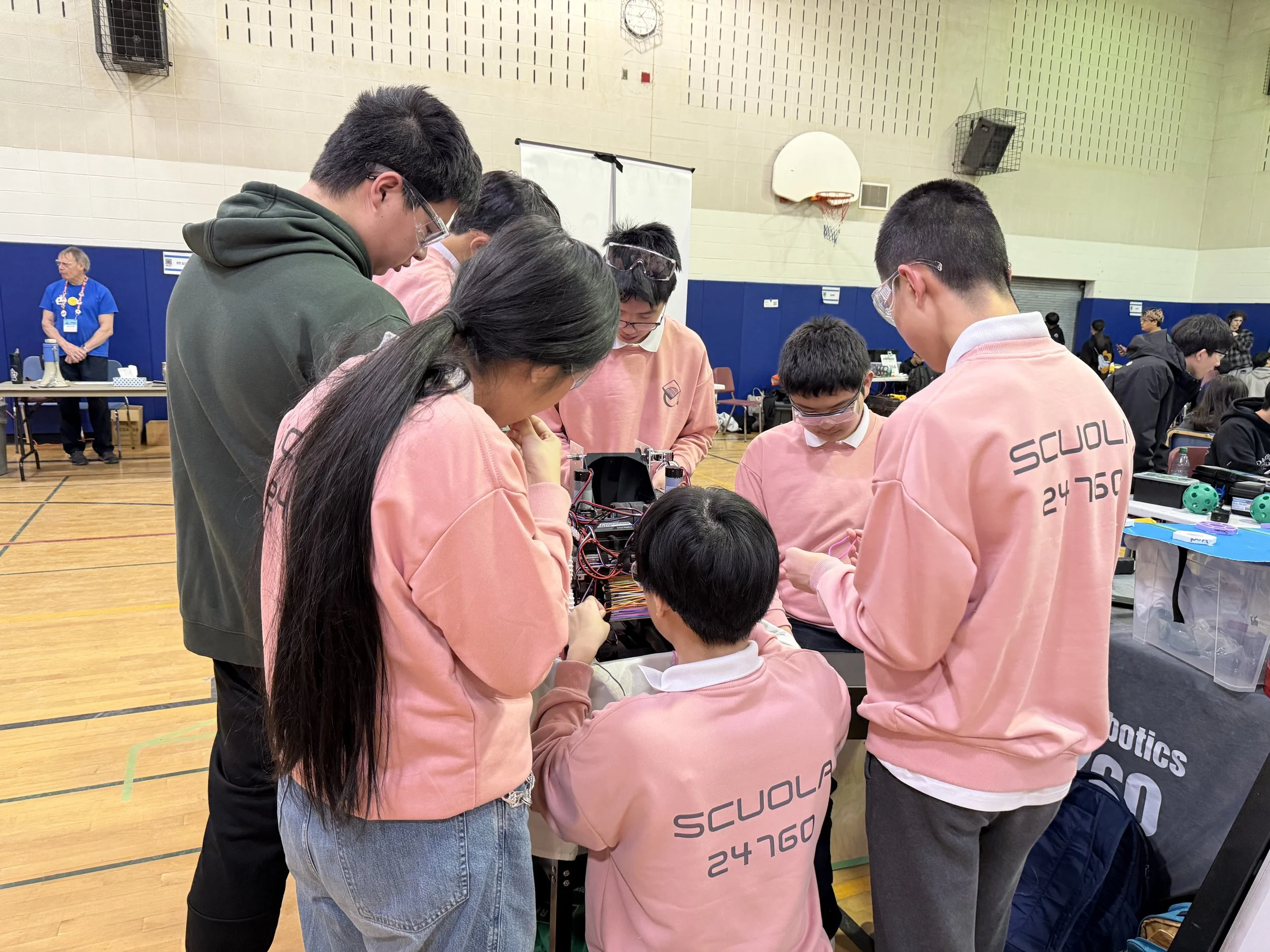 A group of students wearing pink hoodies and safety goggles are gathered around a table working on a robotics project in a gymnasium. There are people in the background, including an older man in a blue shirt and medals, and other students at different tables.