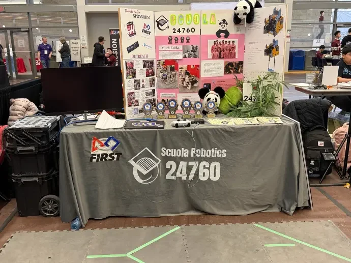 Robotics team display table with a gray tablecloth reading 'Scuola Robotics 24760,' showcasing trophies, panda plush toys, a plant, and a colorful poster board with photos and information, located in an indoor event space.