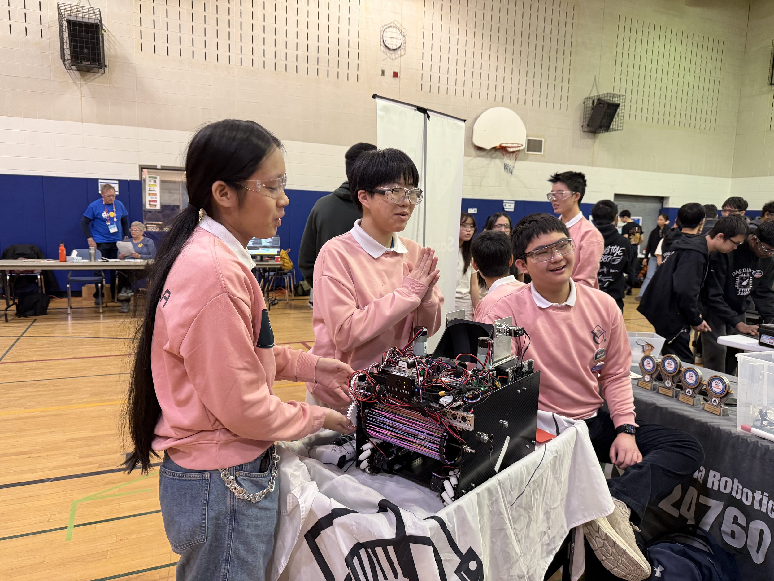 People gathered indoors with some working on robotics projects at a table, wearing pink sweatshirts, surrounded by various electronic components and trophies.