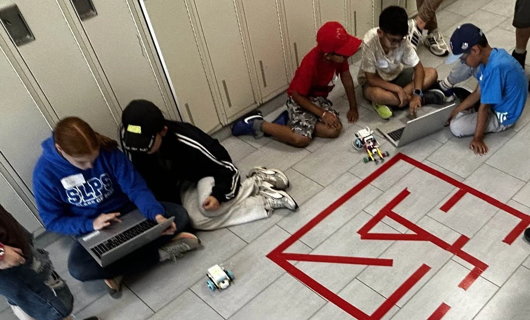 Group of children sit on the floor of a hallway using laptops and working on robots, with a red outlined maze on the tiles spelling out 'KAIS'.