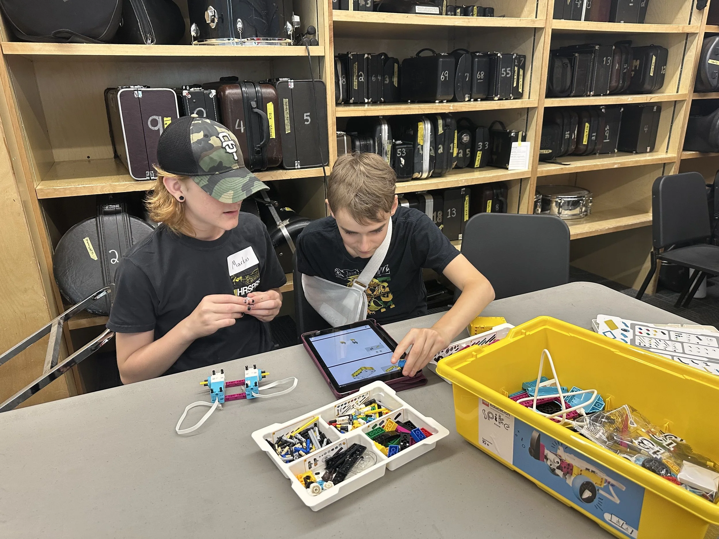 Two children working on a robotics project at a table, with various robot parts and tools in containers. One child is wearing a camouflage baseball cap and a black shirt, while the other is reaching into a tablet. Shelves filled with instrument cases are in the background.