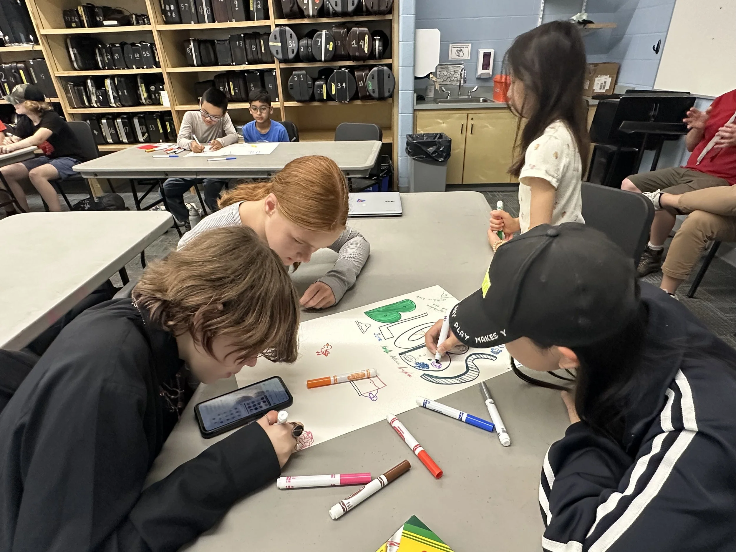 Group of children gathered around a table working on a colorful poster with drawings and writing in a classroom or library setting.