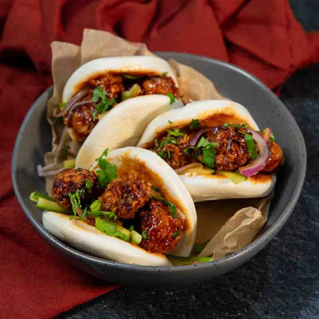 Three Asian-style steamed buns filled with spicy, saucy meat and garnished with chopped cilantro, served in a black bowl.
