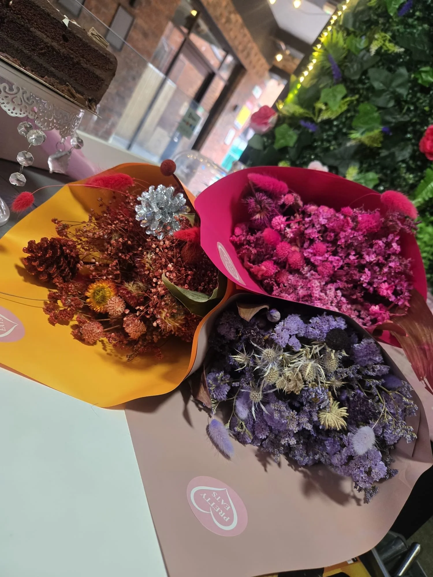 Three brightly colored bouquets of dried flowers arranged on a table at a flower shop, with a pink, yellow, and purple bouquet, and plants visible in the background.