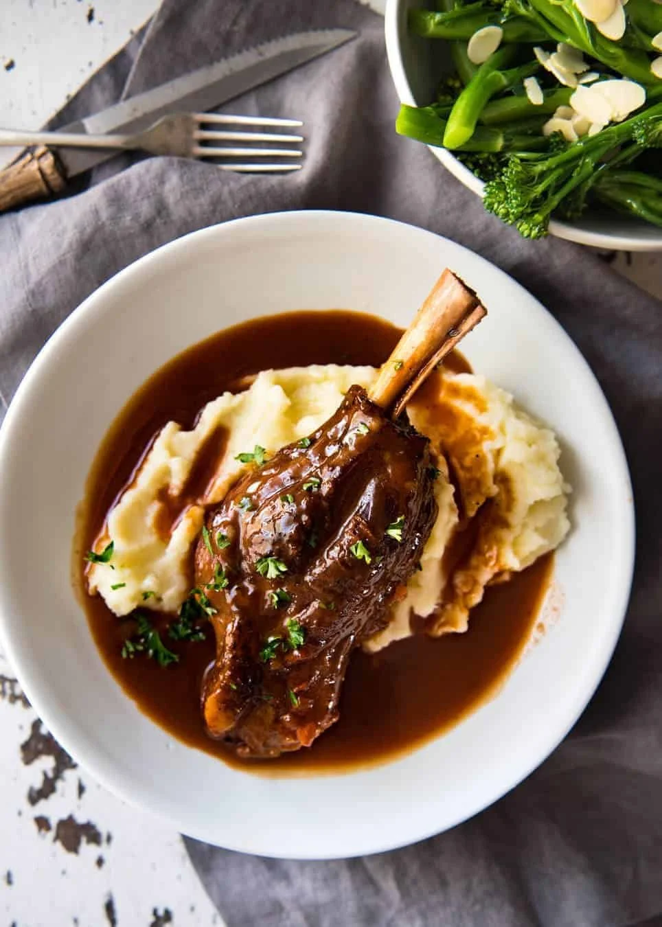 A plate with mashed potatoes topped with a braised lamb shank and gravy, garnished with chopped parsley. To the side is a bowl of cooked green vegetables, possibly broccoli or green beans.