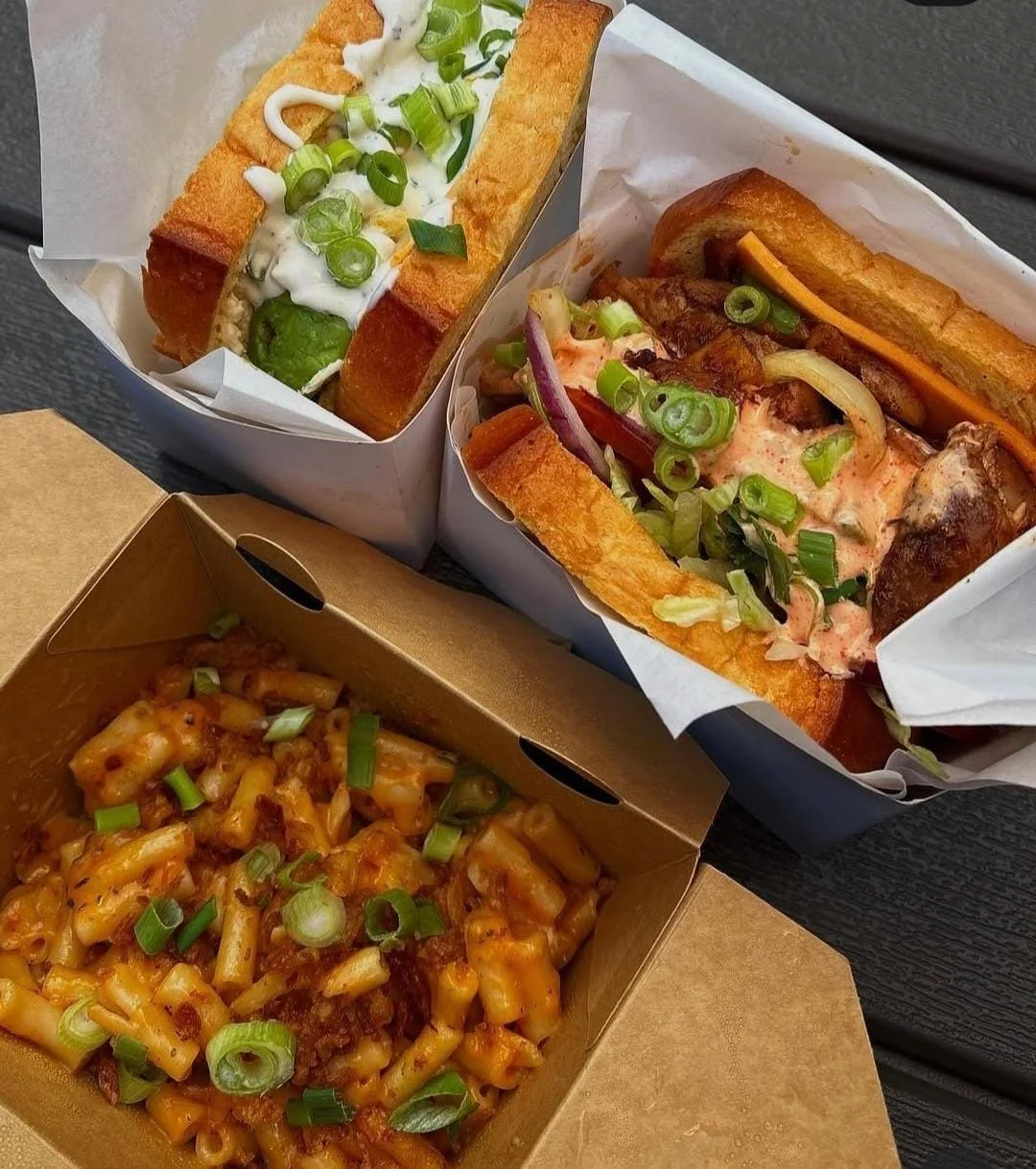 Three takeout containers with different types of loaded hot dogs and macaroni and cheese, topped with chopped green onions.