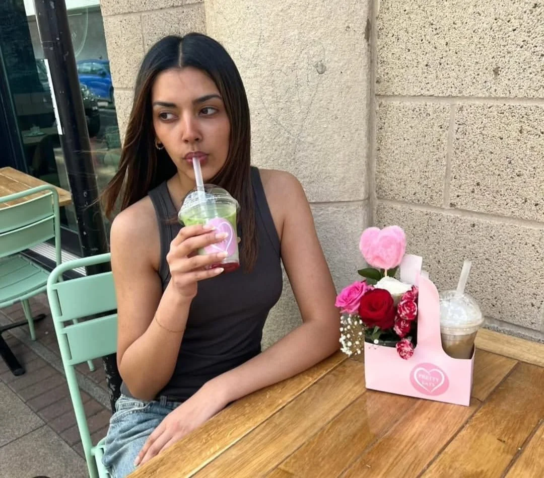 Young woman sitting at an outdoor table, drinking a frappuccino, with a pink bouquet of flowers and a pink love heart decor in a small pink box on her right.