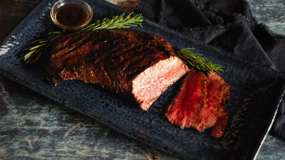 Barbecued beef brisket with a slice cut, served on a black plate with rosemary sprigs and a small cup of barbecue sauce.