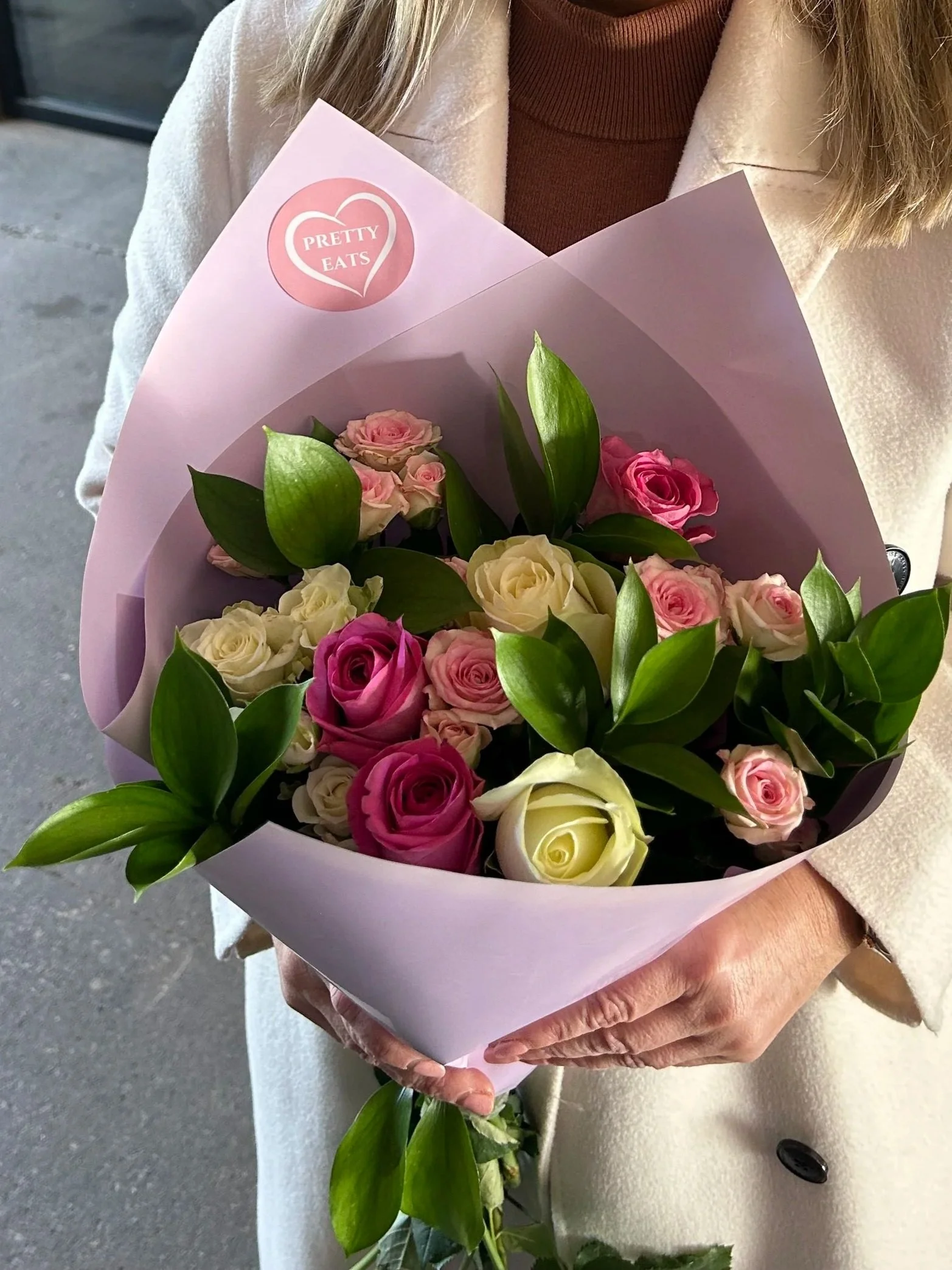 A woman holding a bouquet of pink, cream, and white roses with greenery wrapped in light pink paper, with a sticker that says "Pretty Eats".