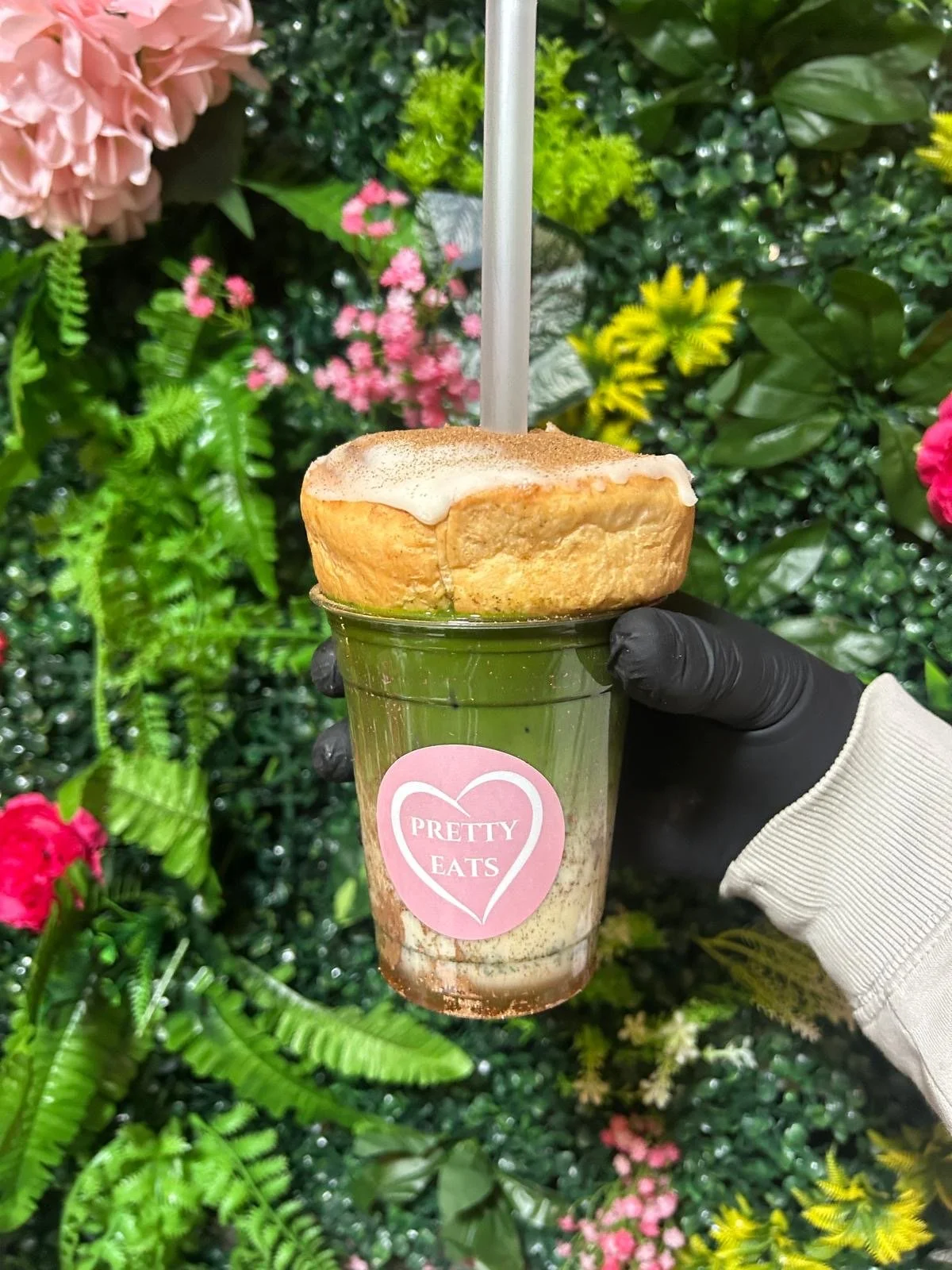 A hand wearing a black glove holds a frappuccino with a pink sticker that says 'Pretty Eats.' The drink is topped with a large, round donut with white icing and cinnamon. The background features a lush, colorful floral display.
