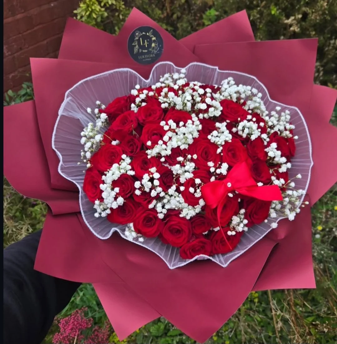 A bouquet of red roses with white baby's breath flowers wrapped in pink and white paper, featuring a pink bow. There is a small black circular logo with gold accents on top of the bouquet.