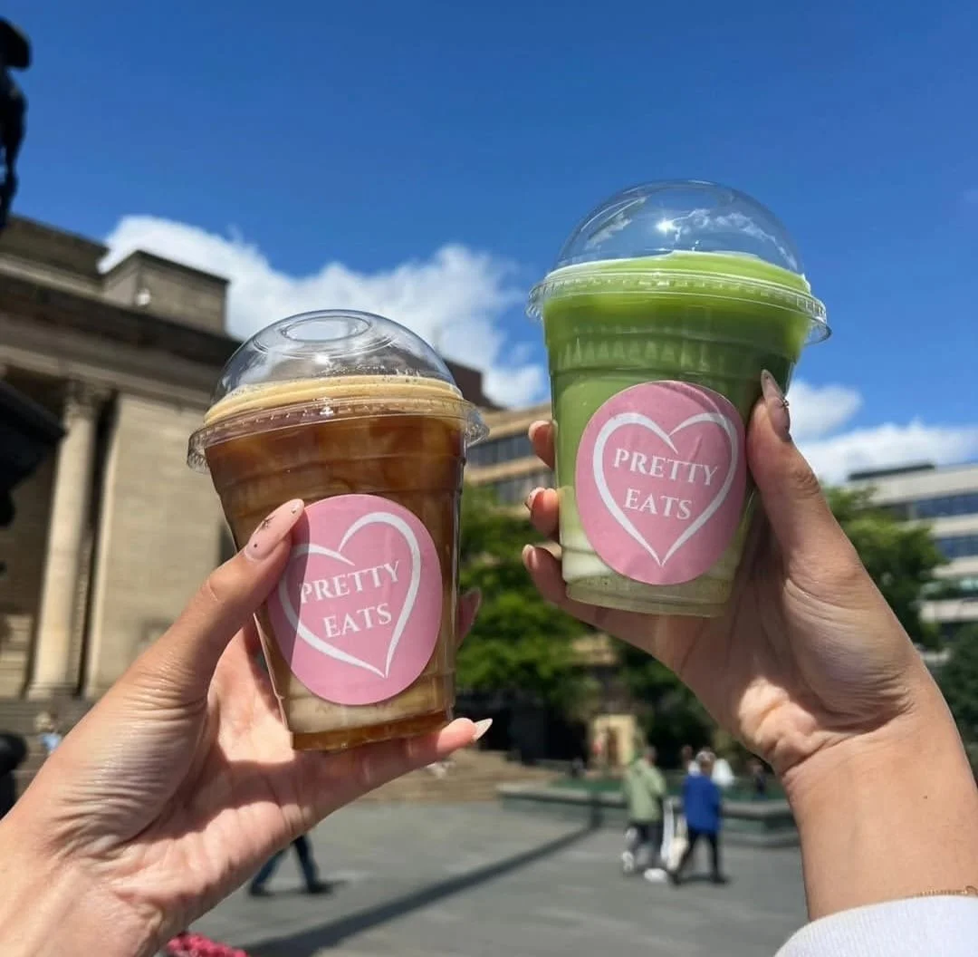 Two hands holding plastic cups with domed lids, one with iced coffee and the other with iced matcha, both with pink stickers that say 'PRETTY EATS' in a heart-shaped design, outdoors on a sunny day with a clear blue sky and people in the background.